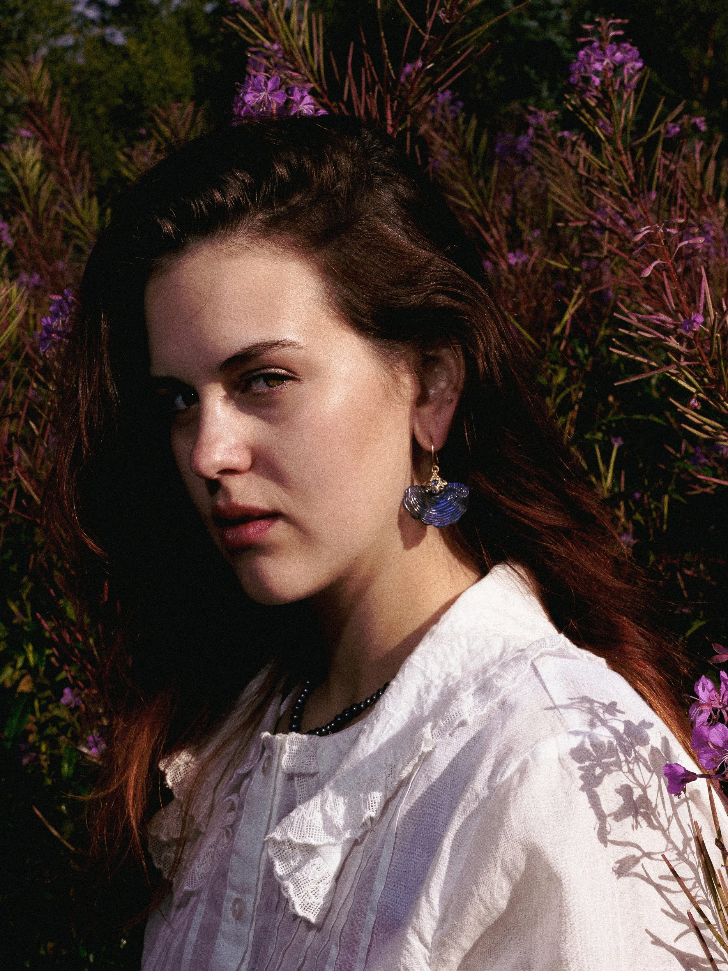 A young woman with dark brown hair and earrings stands amidst purple flowers, wearing a white blouse with lace detailing.