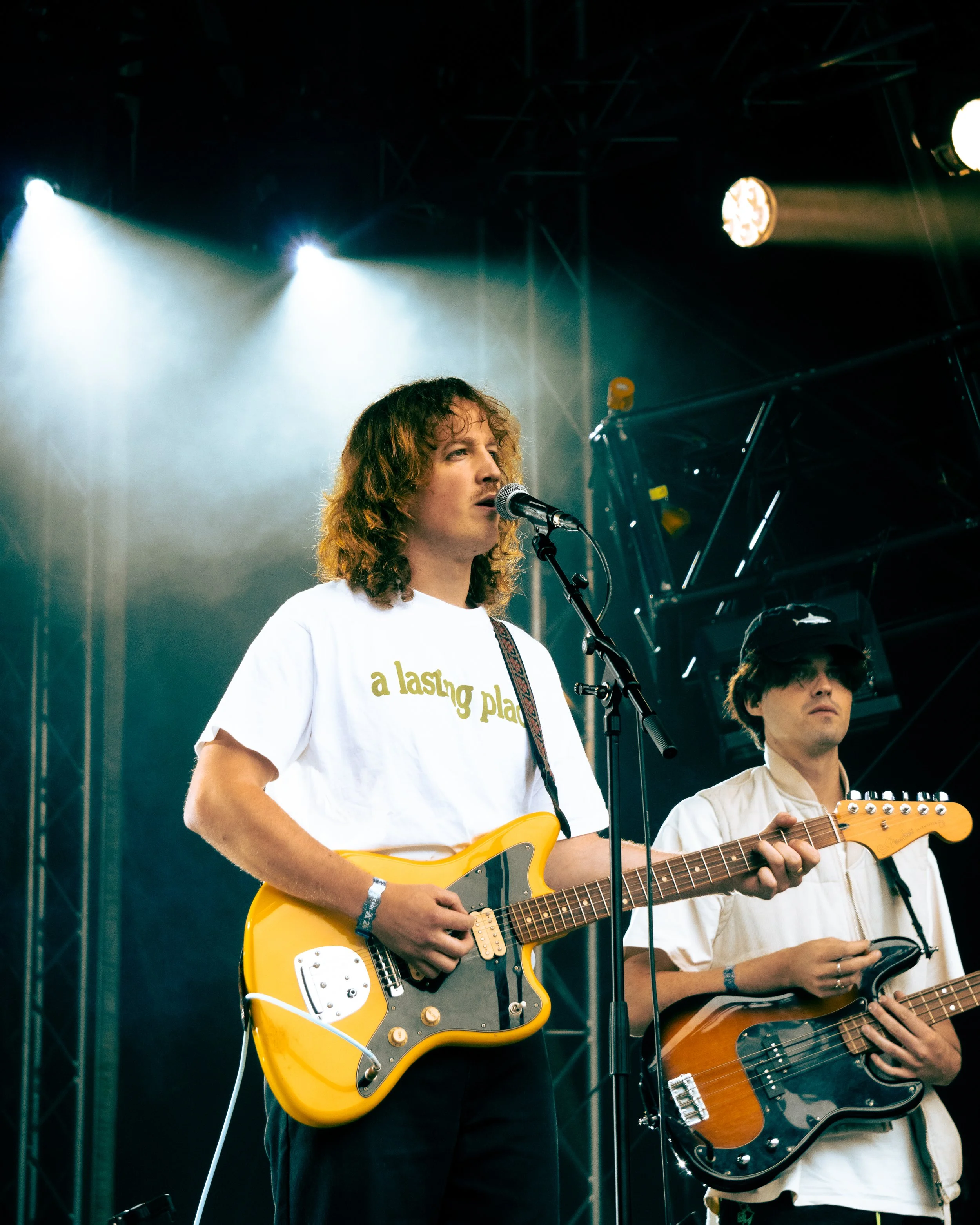 Two musicians perform on stage, one playing yellow electric guitar and singing into a microphone, the other playing bass guitar. Stage lights shine down.