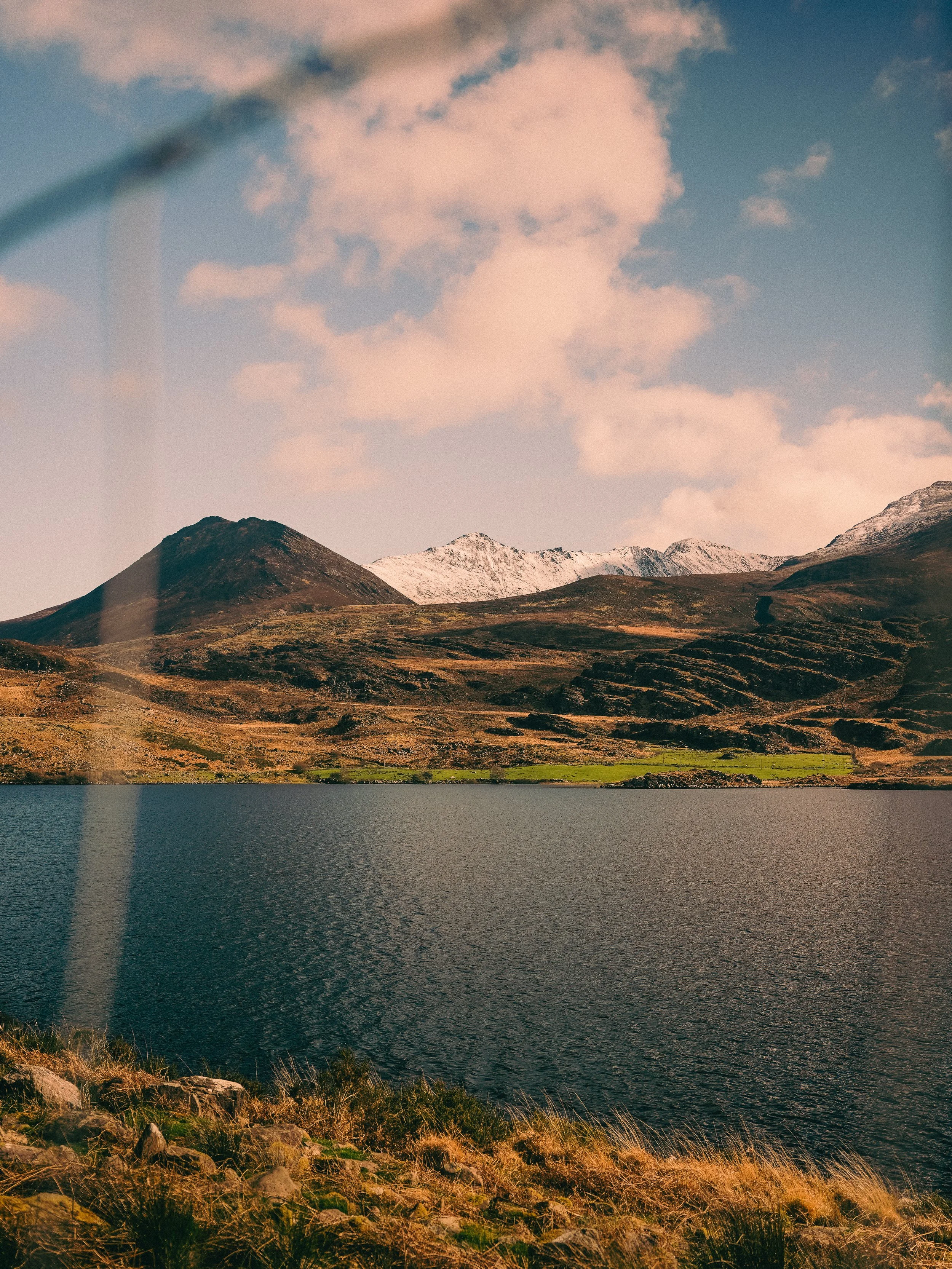 Scenic landscape of mountains with snow caps, a lake in the foreground, and grassy terrain along the shoreline under partly cloudy sky.