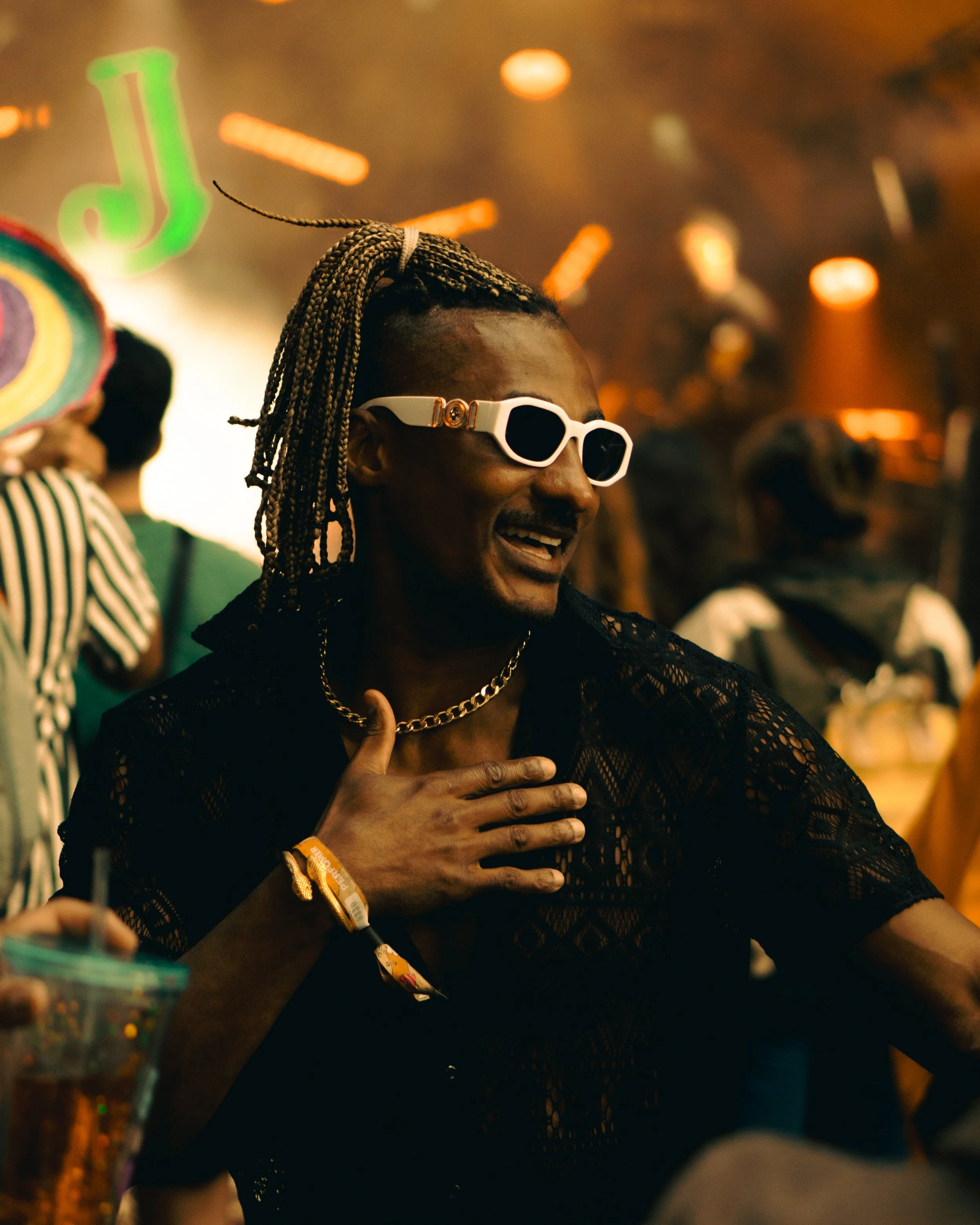 A man with braided hair wearing white sunglasses, a black lace shirt, and a gold chain, smiling and touching his chest with his hand in a lively party setting.