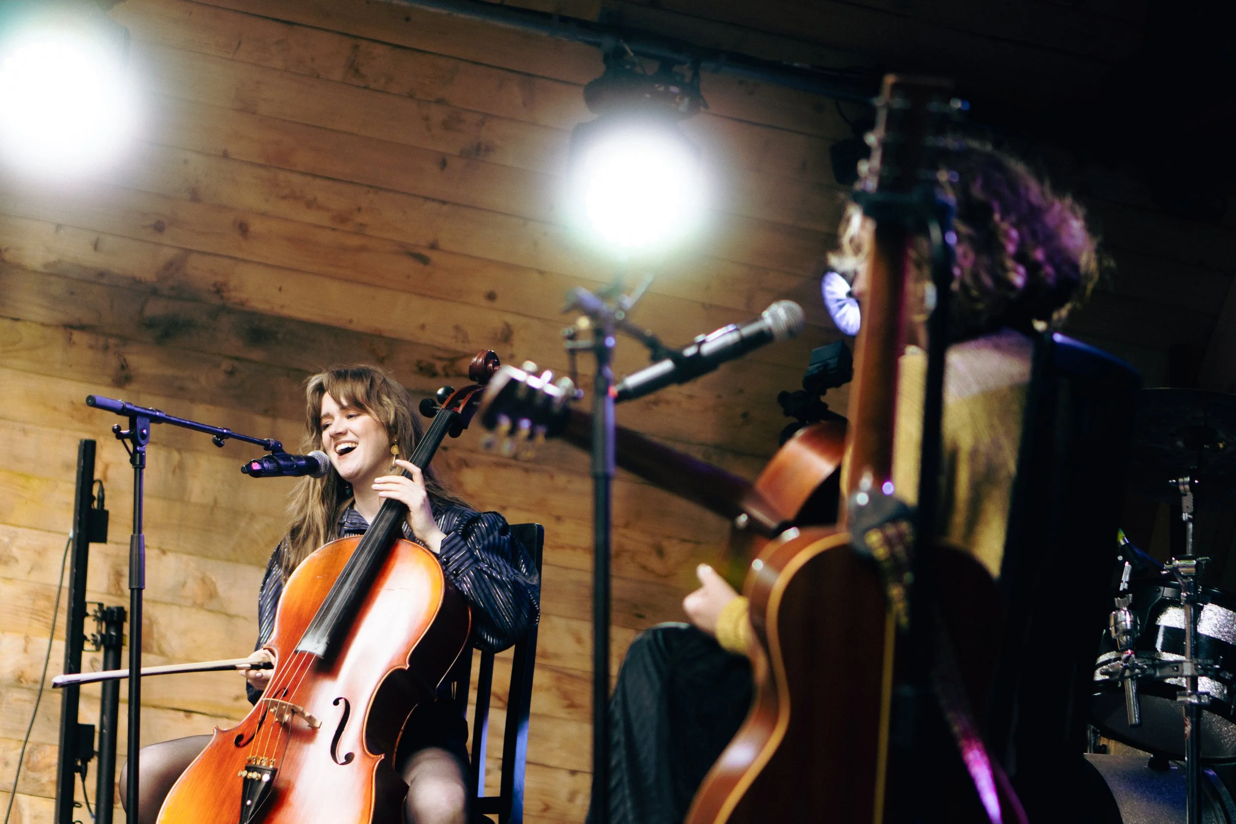 A young woman playing a cello, smiling and singing into a microphone, in a wooden music venue, with another musician partially visible playing a guitar.