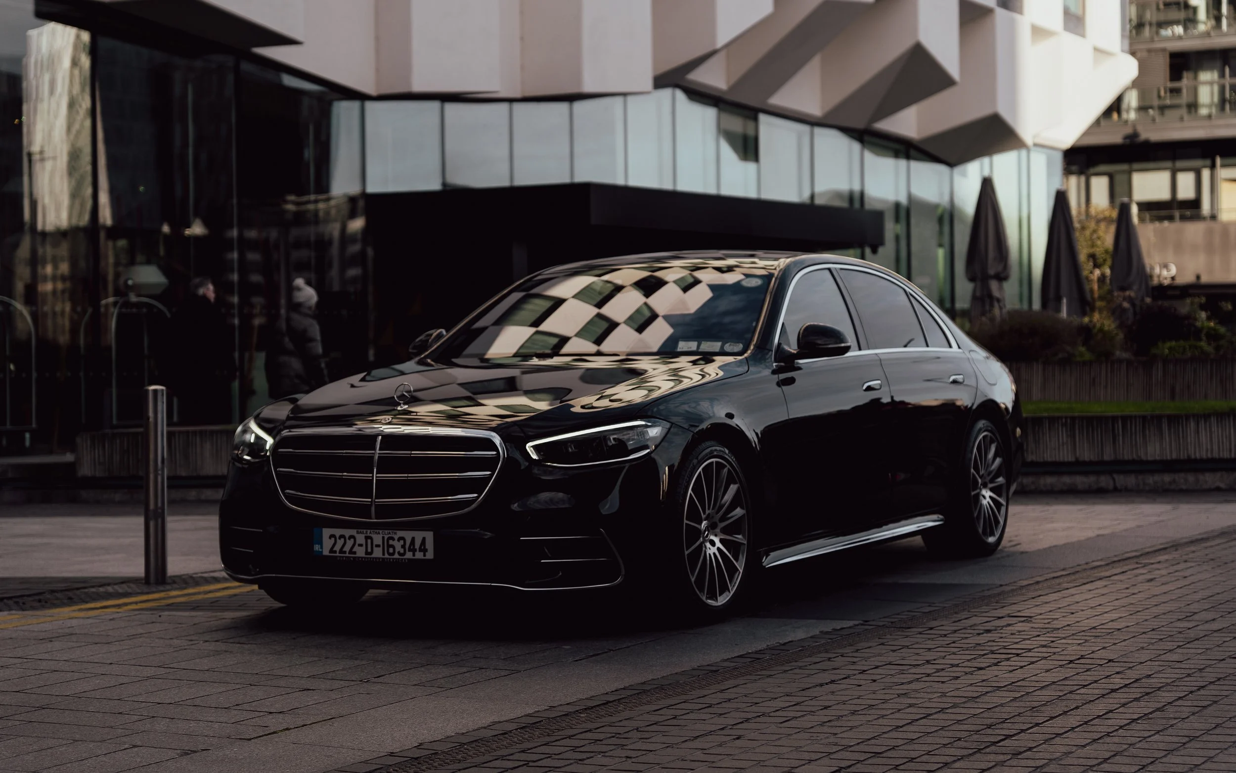 A black luxury sedan, Mercedes-Benz, parked on a city street in front of a modern glass building with reflective windows.