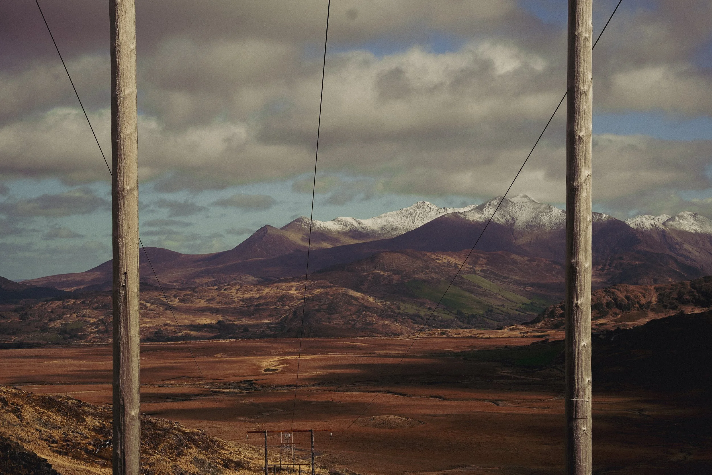 A landscape view of mountains with snow-capped peaks, rolling hills, a valley, and electrical poles in the foreground under a cloudy sky.