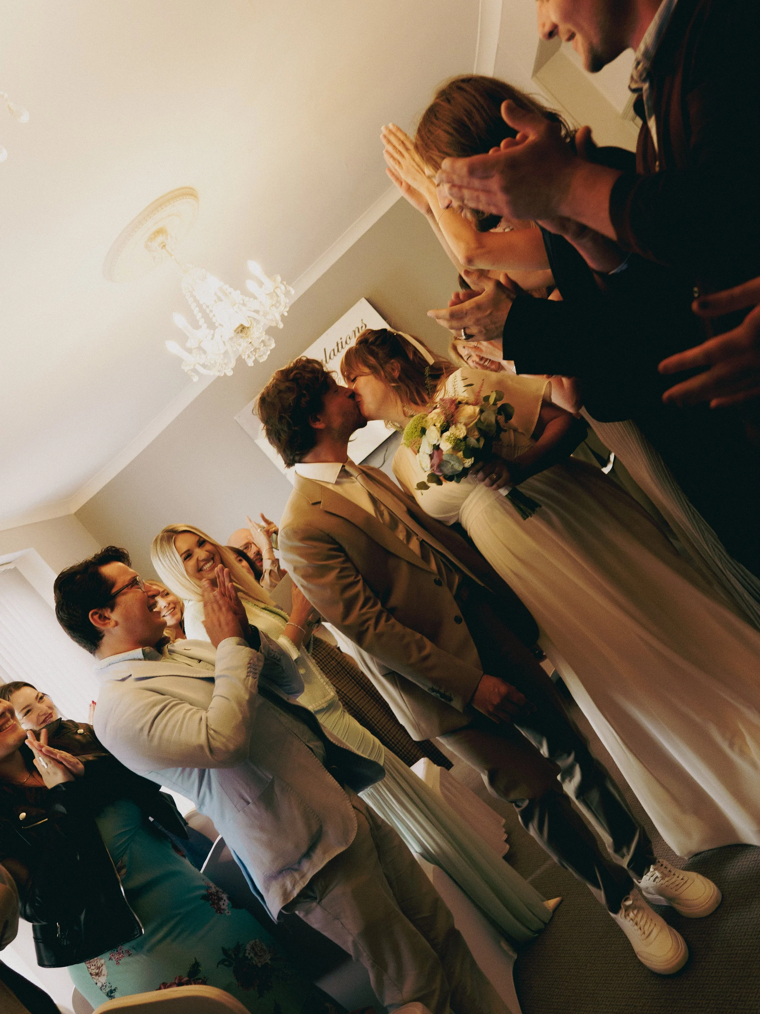A group of people at a wedding ceremony, with a bride and groom kissing, surrounded by guests cheering and clapping in an indoor setting with a chandelier and a sign that reads 'National' in the background.