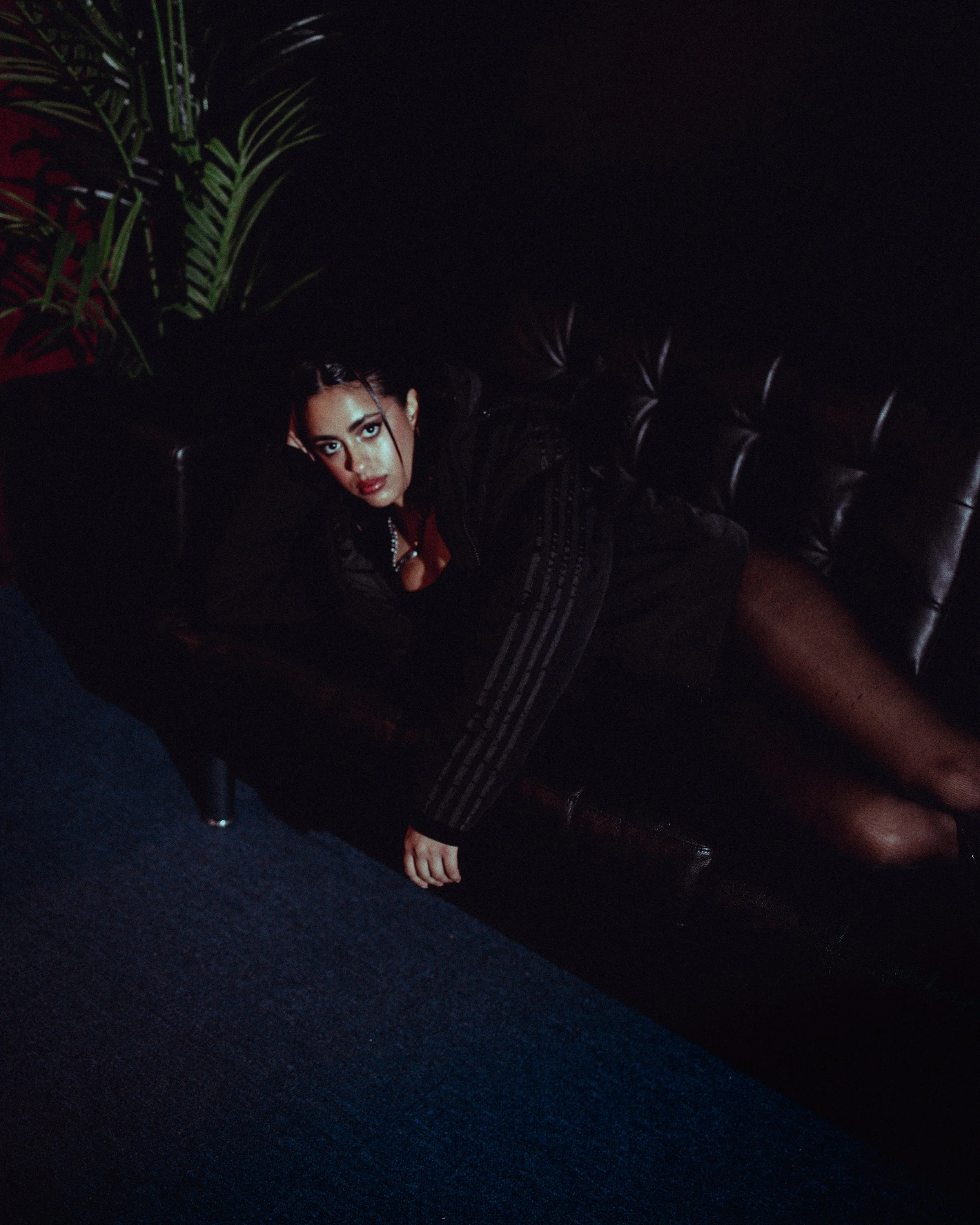 A young woman with dark hair and makeup lying on a black leather couch in a dimly lit room, with a green plant in the background.