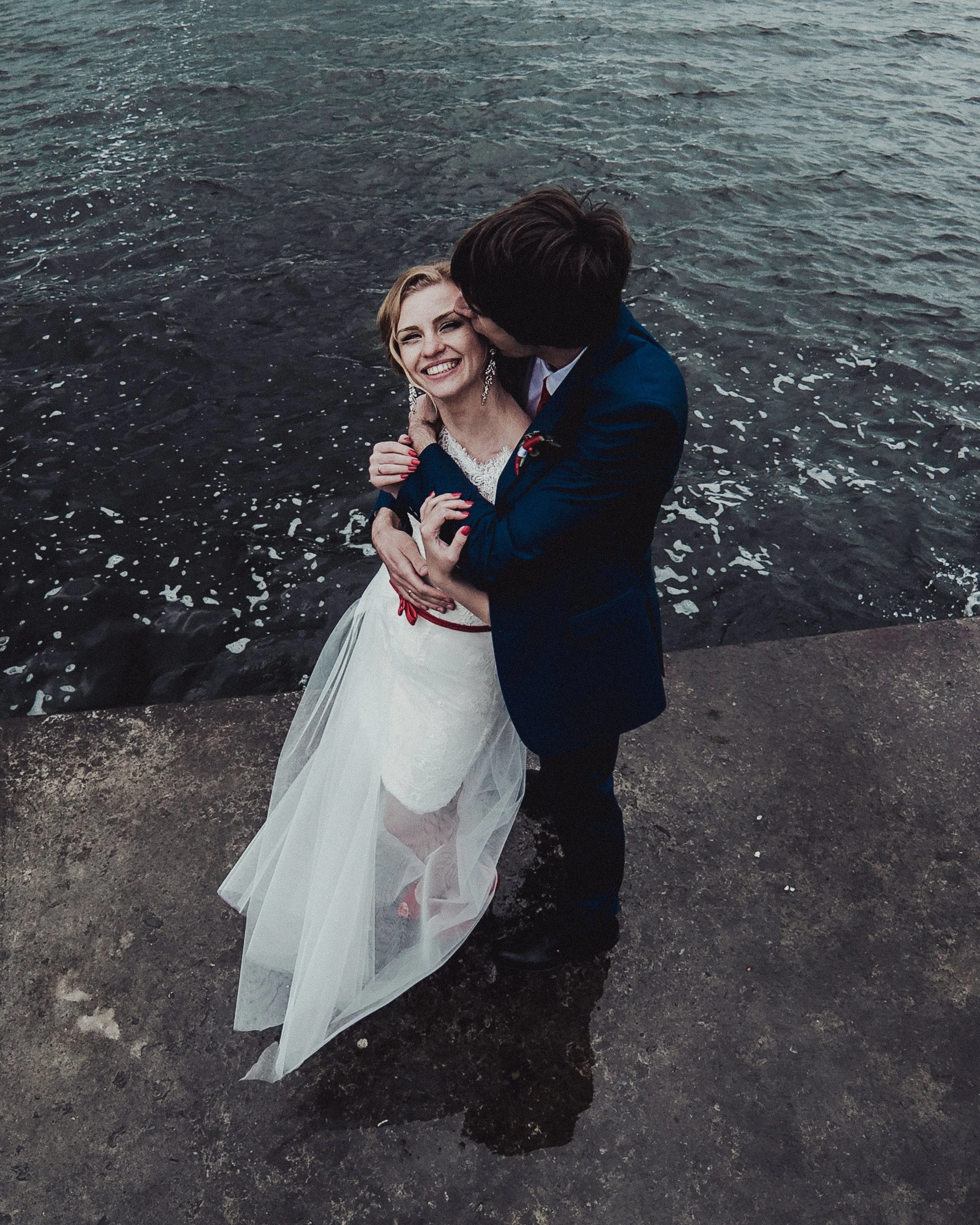 A bride and groom standing on a concrete surface by the water, sharing a moment of affection during their wedding, with the groom kissing the bride on the cheek.