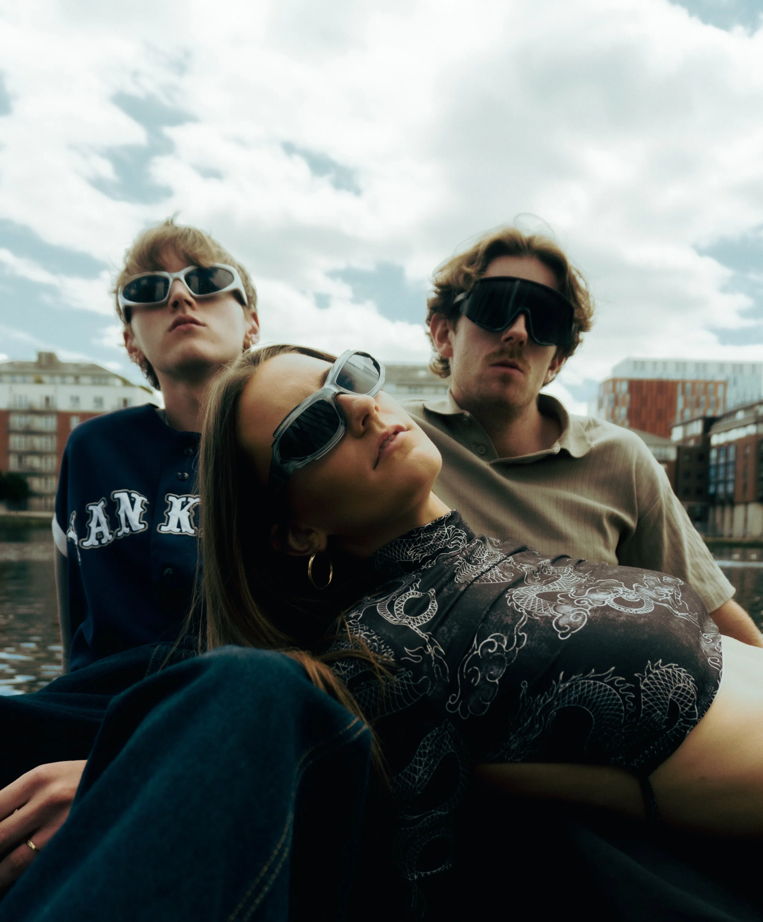 Three young adults wearing sunglasses relax outdoors by a body of water, with modern buildings in the background under a cloudy sky.