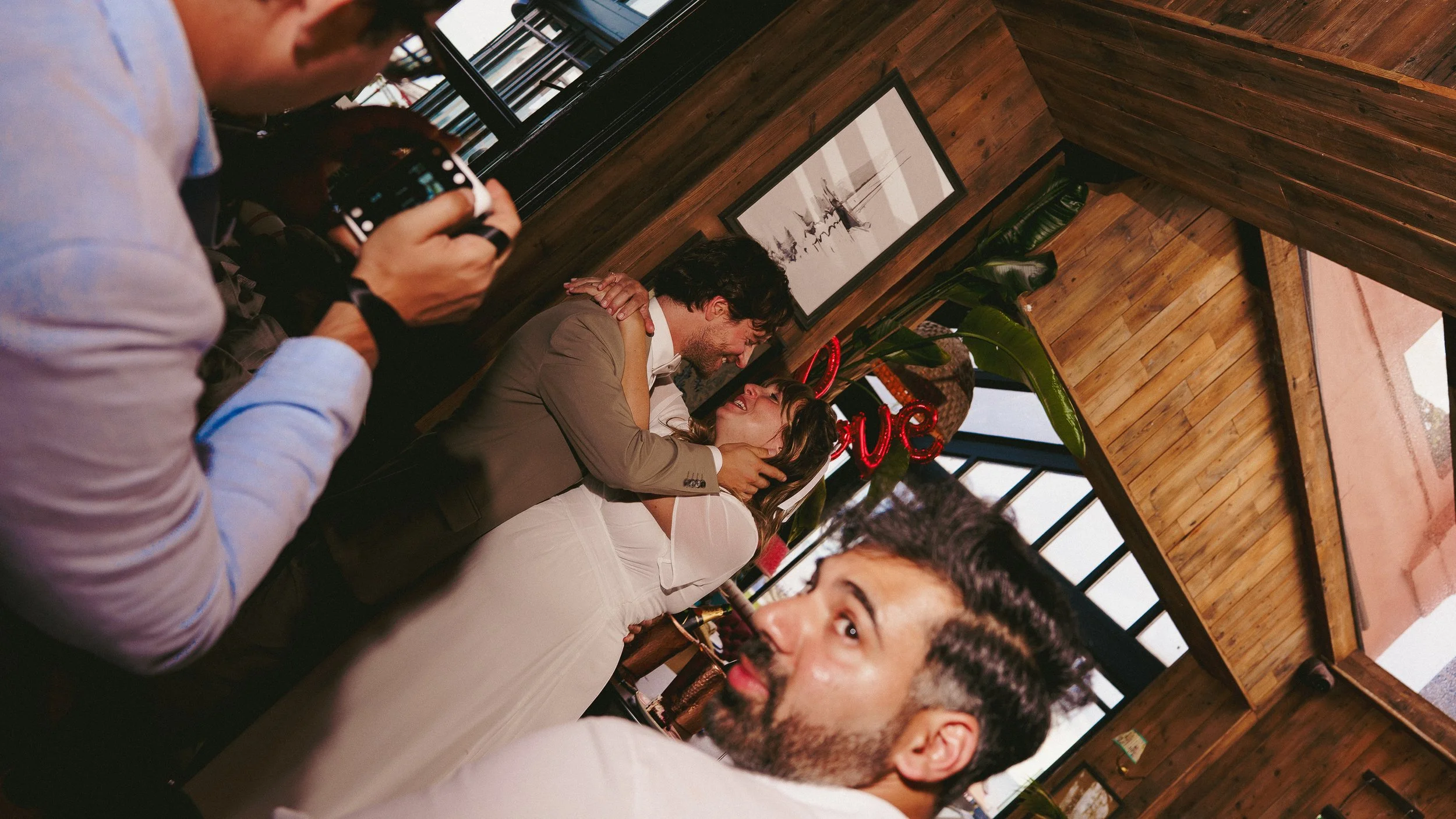 A group of people dancing and enjoying a celebration inside a wooden-paneled room with a neon "Love" sign and framed artwork on the wall.