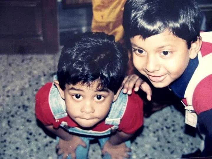 Two young boys leaning forward and smiling at the camera inside a room.