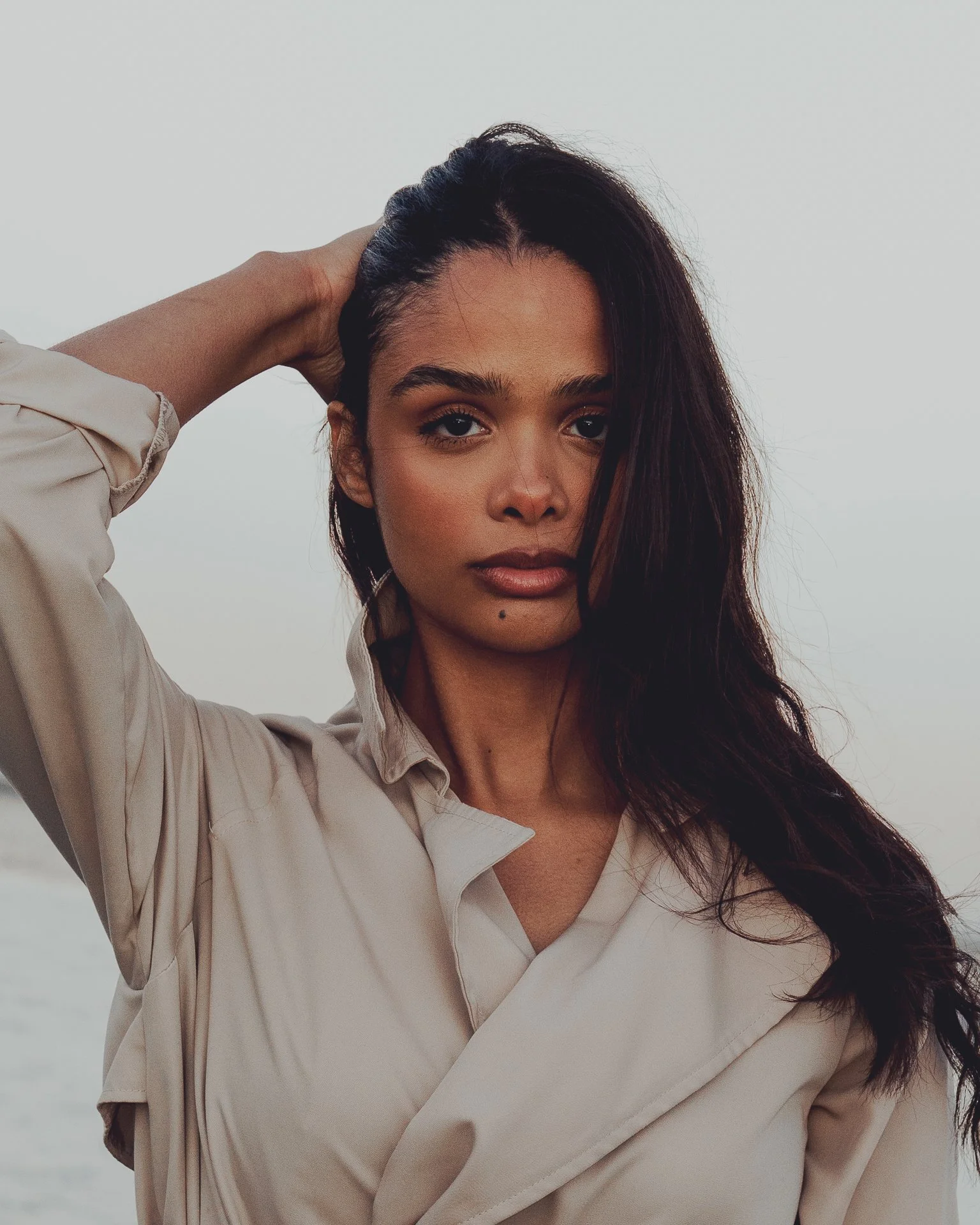 A woman with long dark hair and tan skin standing outdoors, with her right hand resting on the back of her head, wearing a beige shirt, with an overcast sky in the background.