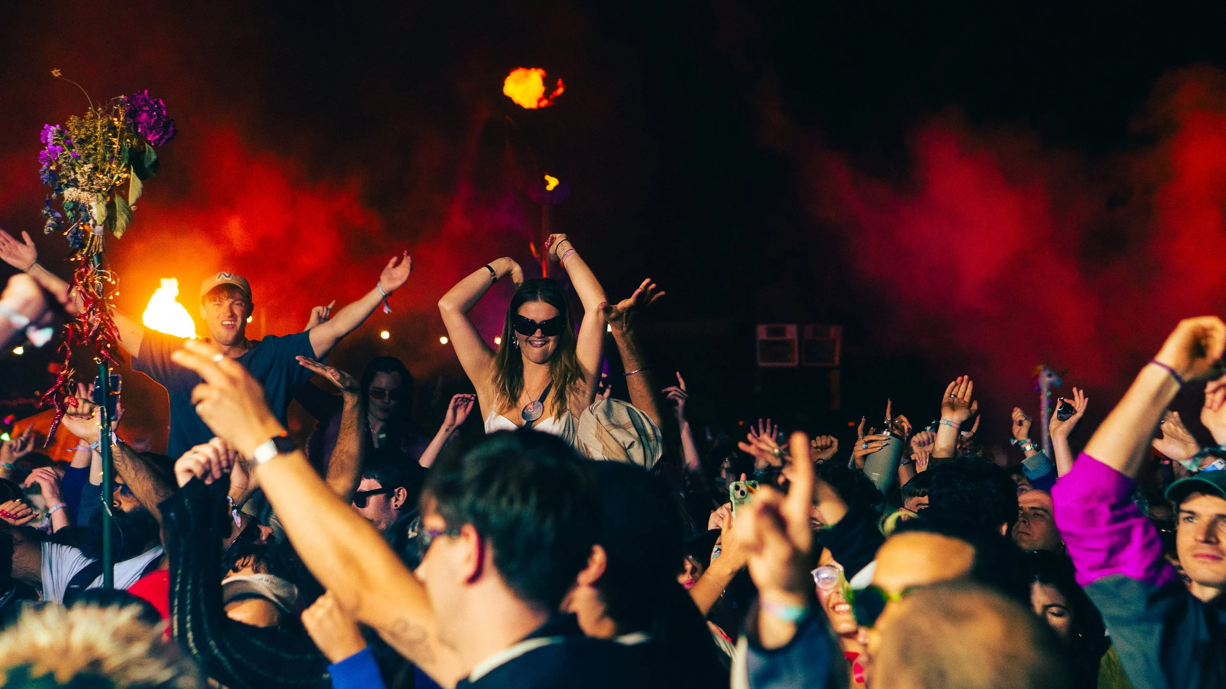 Crowd of people dancing and celebrating at night with colorful lights and smoke, some raising their hands.