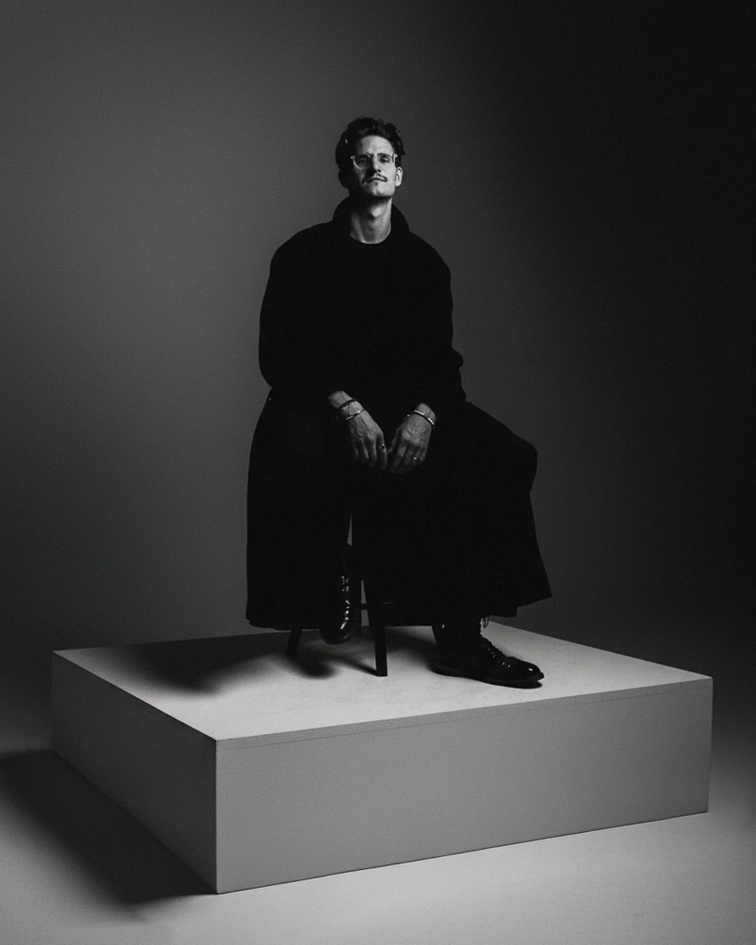 Black and white photo of a man sitting on a chair on a raised platform, wearing dark clothing, with his hands resting on his lap, against a plain background.