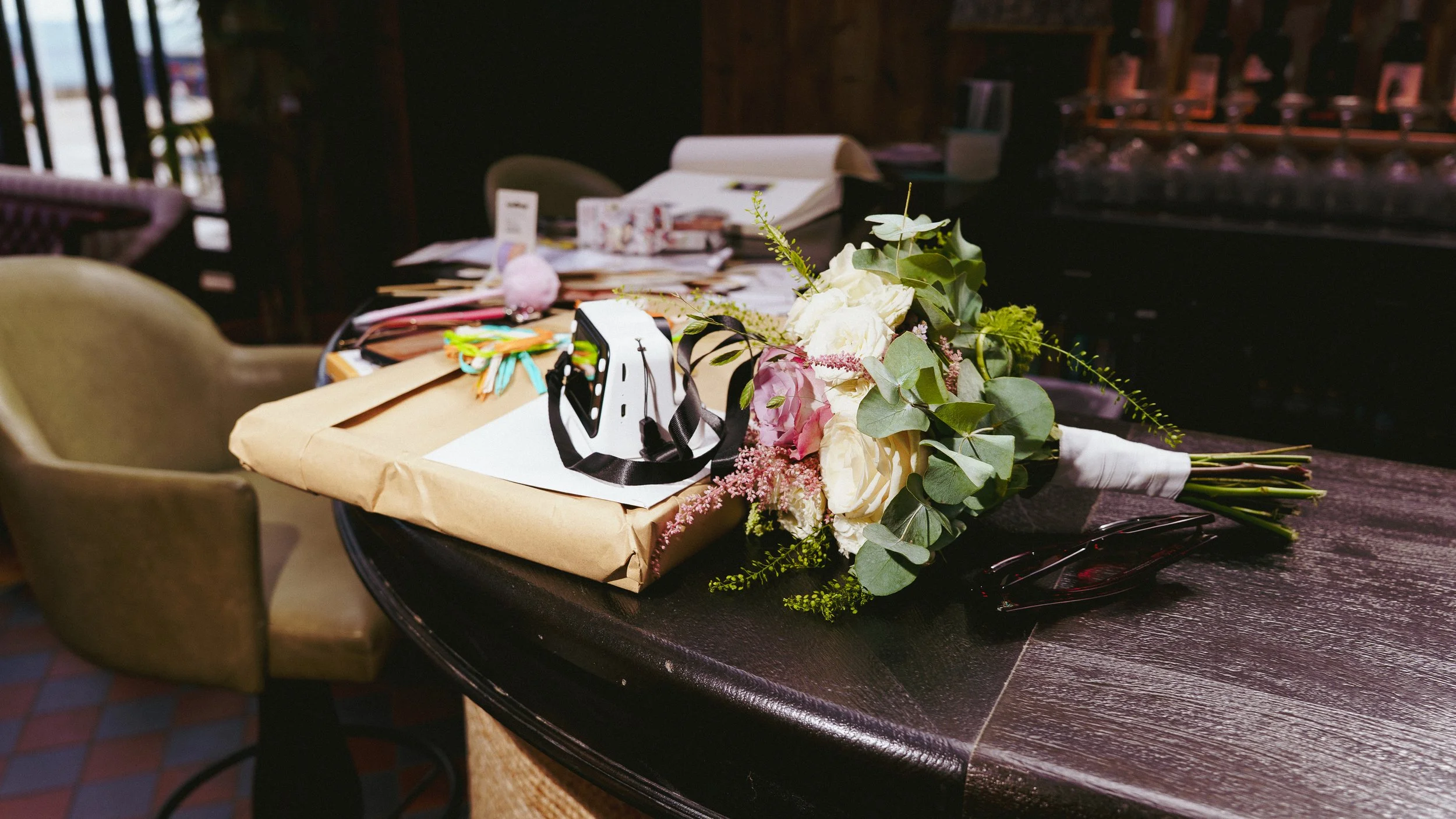 A bouquet of white and pink flowers with greenery, a gift wrapped in brown paper, around  the flowers and gift are colorful ribbons and a white paper, and a pair of sunglasses on a dark wooden table.