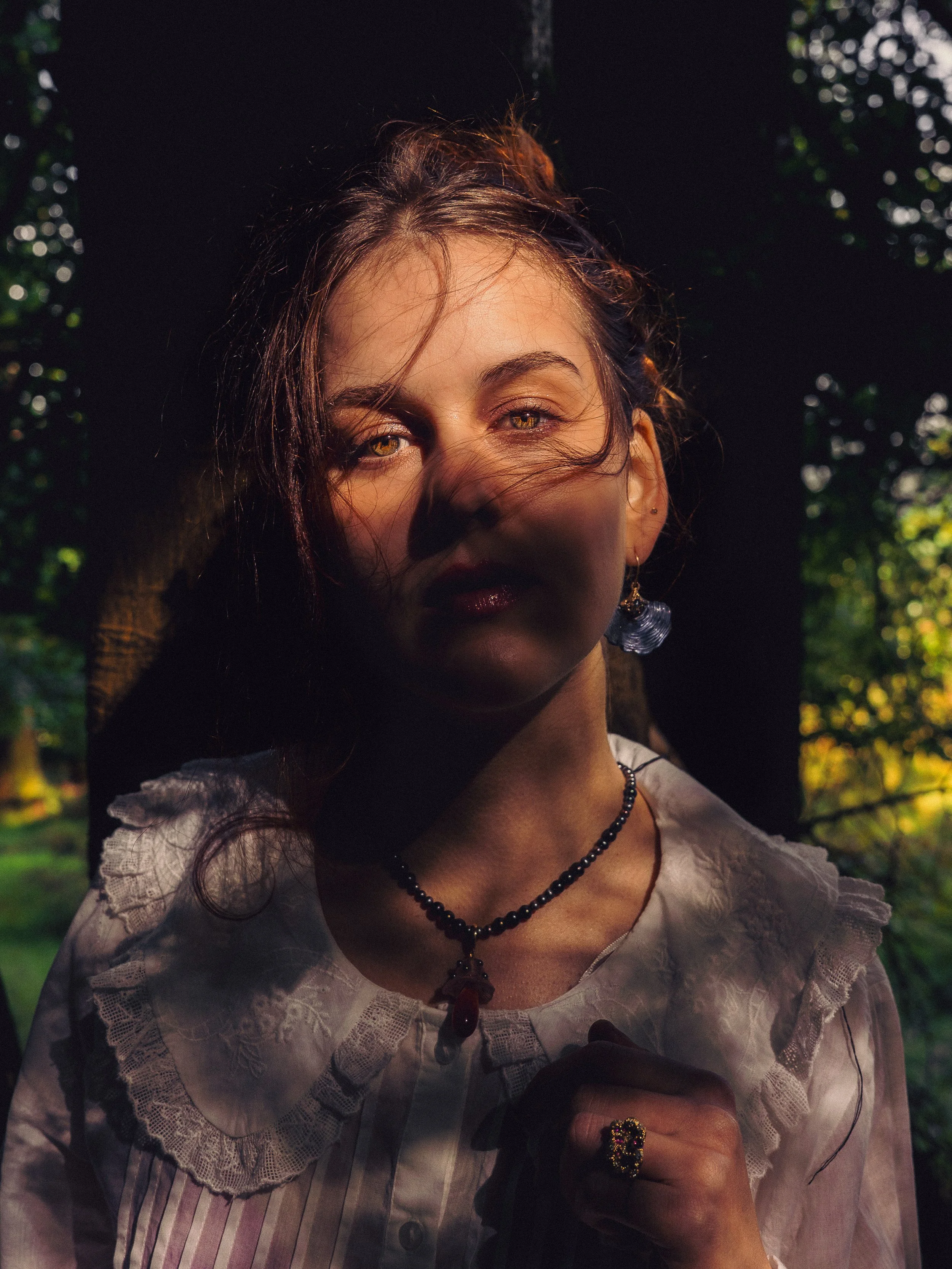 A woman with light skin and dark hair standing outdoors in a sunlit forest, partially shadowed by a tree, wearing a white lace-collared blouse, jewelry, and looking at the camera.