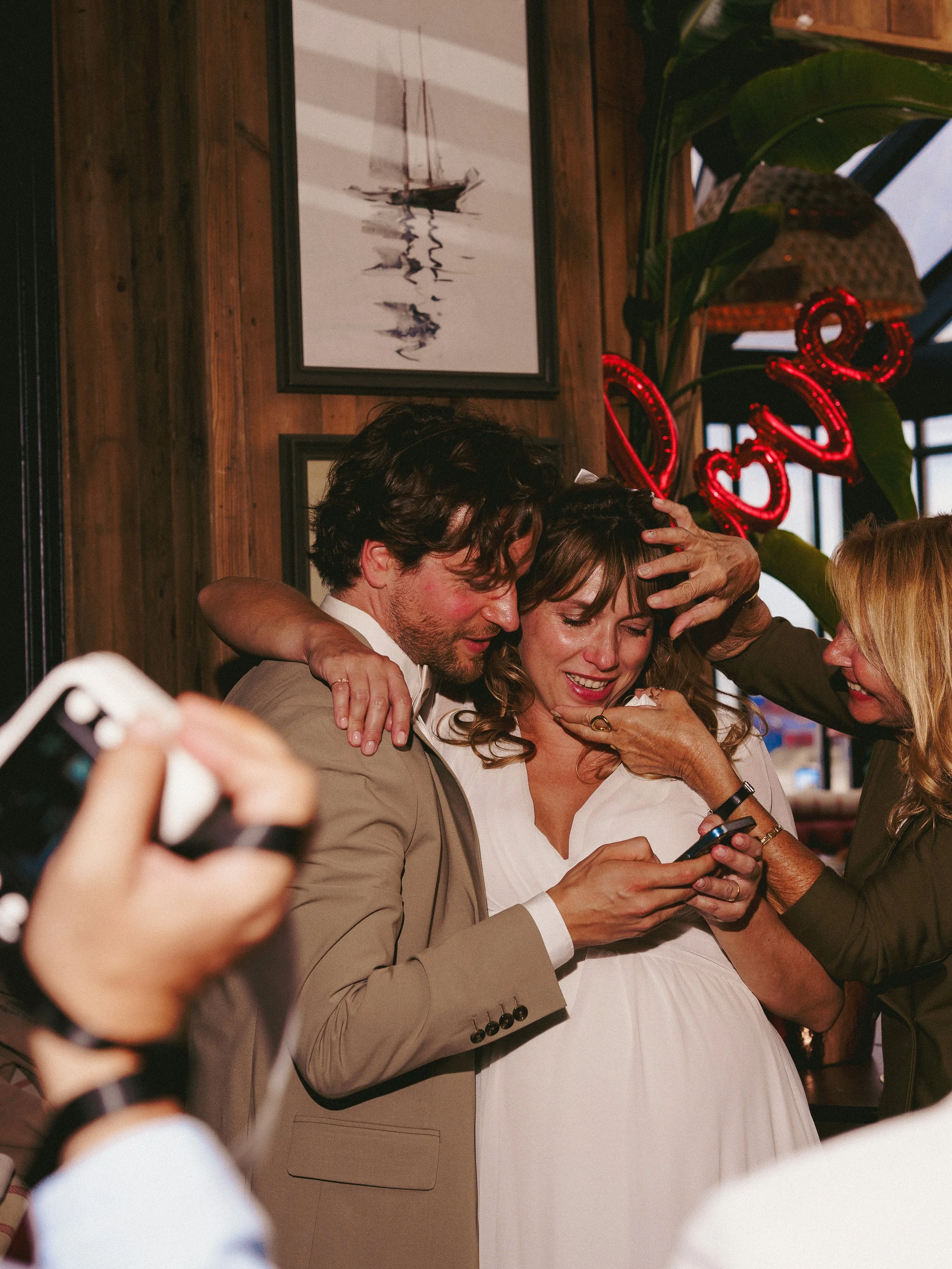 People celebrating at a party, with one woman in a white dress appearing pregnant, surrounded by others, some holding phones, indoors with wooden walls and artwork.