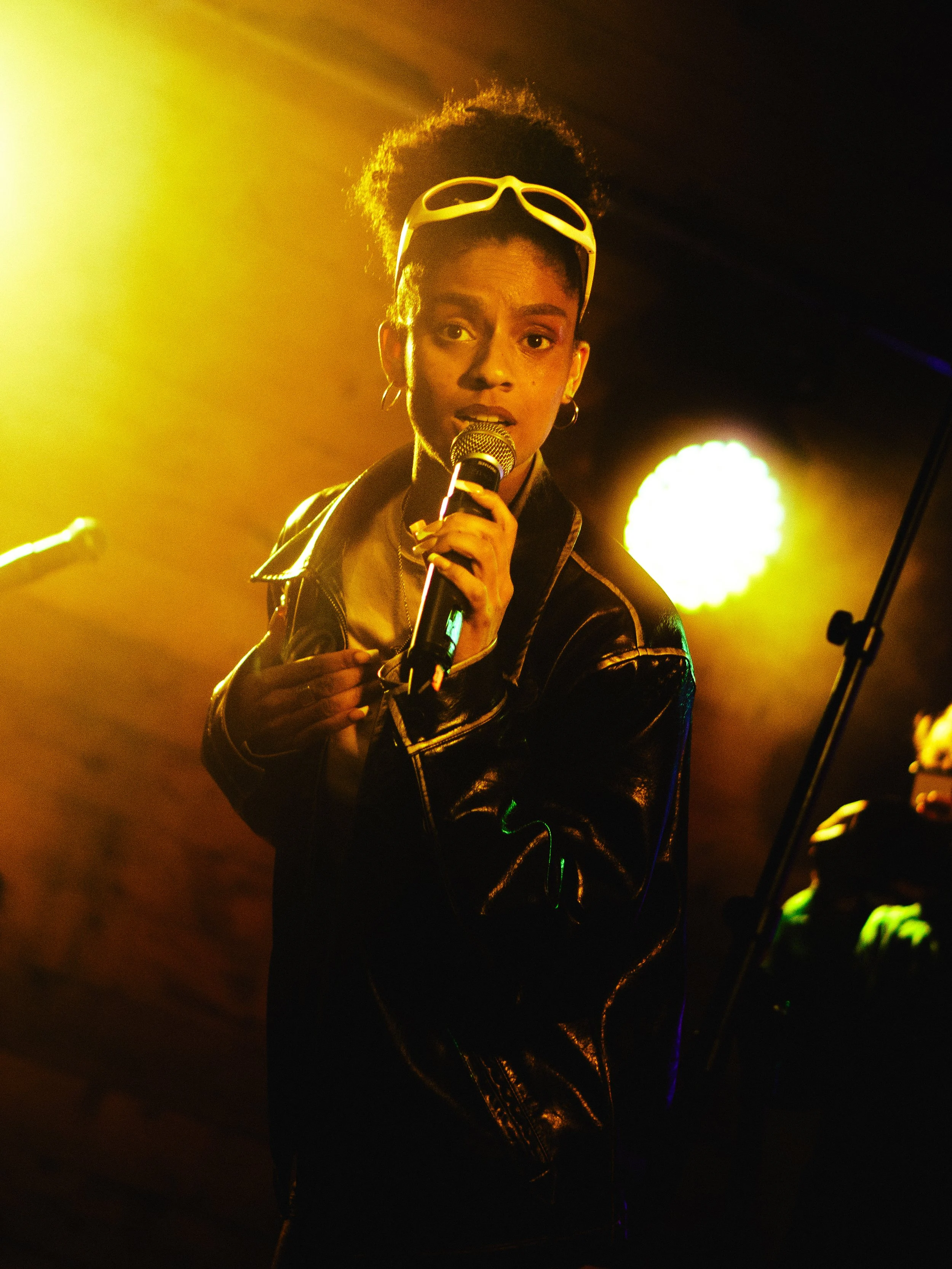 A woman singing on stage with gold and black lighting, wearing a black leather jacket and white sunglasses on her head.