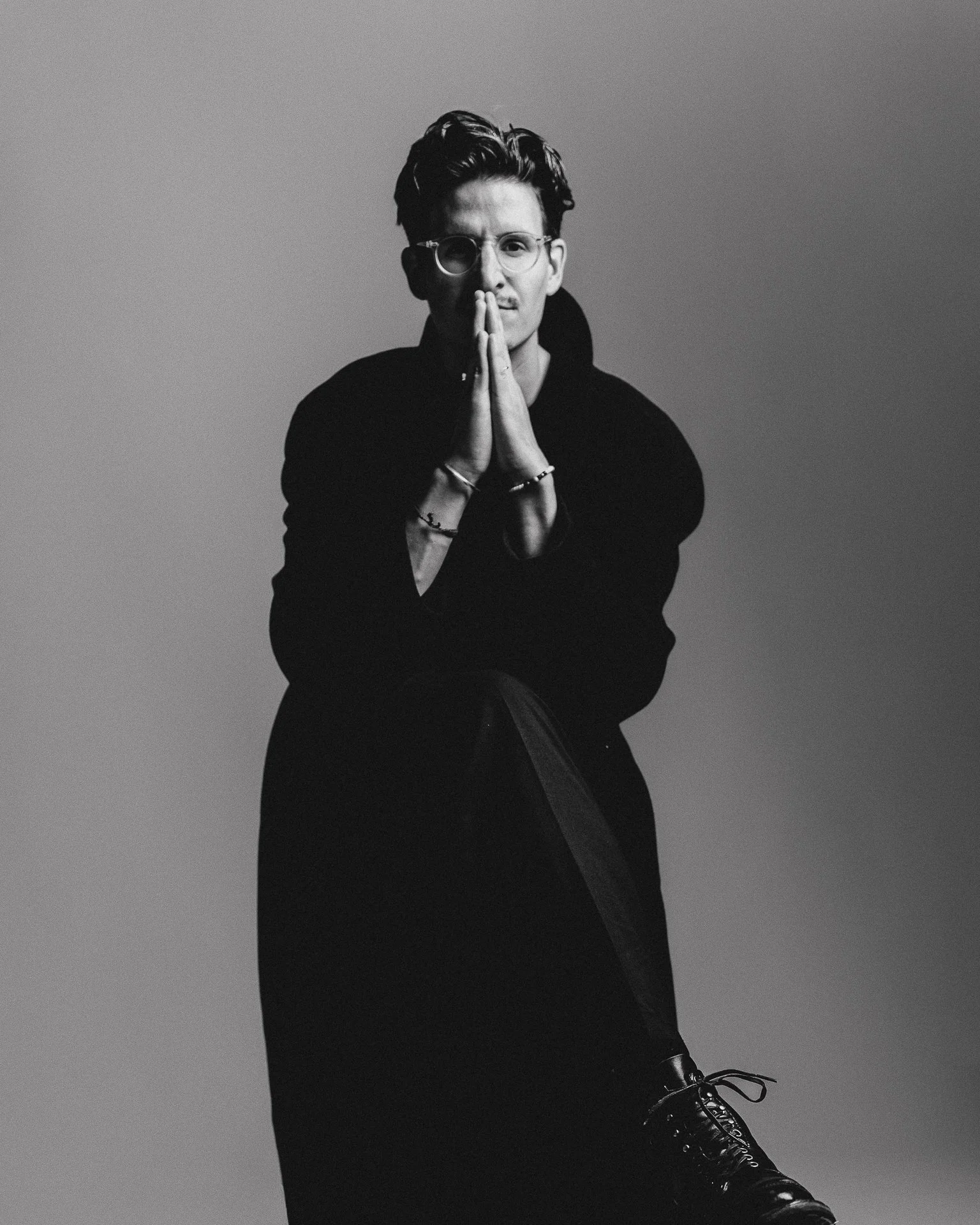A black and white photo of a young man with glasses, wearing a dark hoodie, kneeling with hands pressed together in a prayer-like gesture, against a plain background.