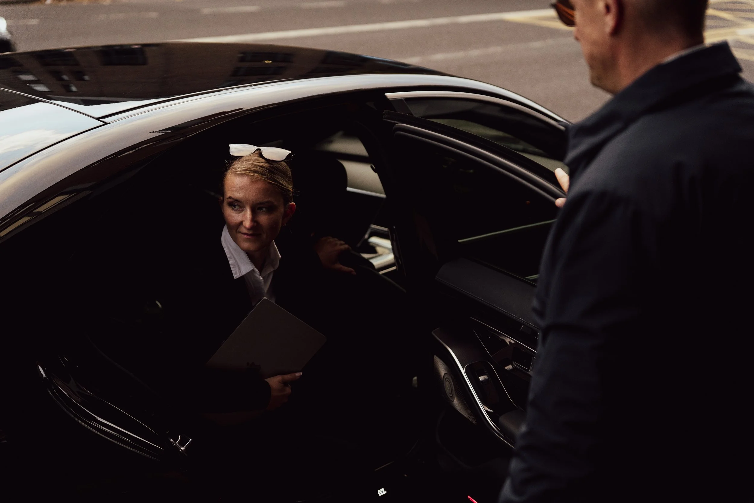 A woman sitting inside a black car, looking up at a man standing outside near the open car door. The woman has blonde hair, sunglasses on her head, and is holding a tablet. The man is wearing a dark jacket and is smiling.