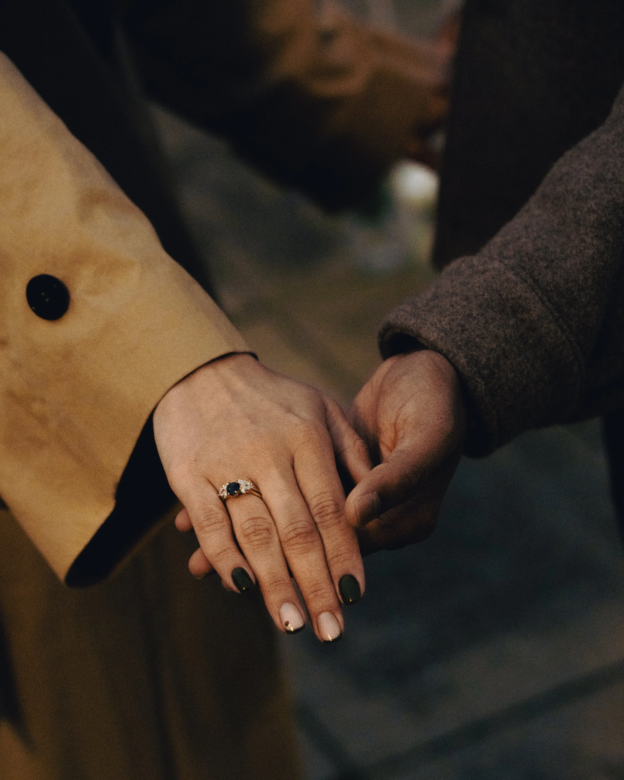 A close-up of a couple holding hands, with the woman's hand resting on top of the man's. The woman is wearing a gold ring with a dark gemstone and has dark green and light pink nail polish. The man is wearing a brown coat and a dark gray or black sle