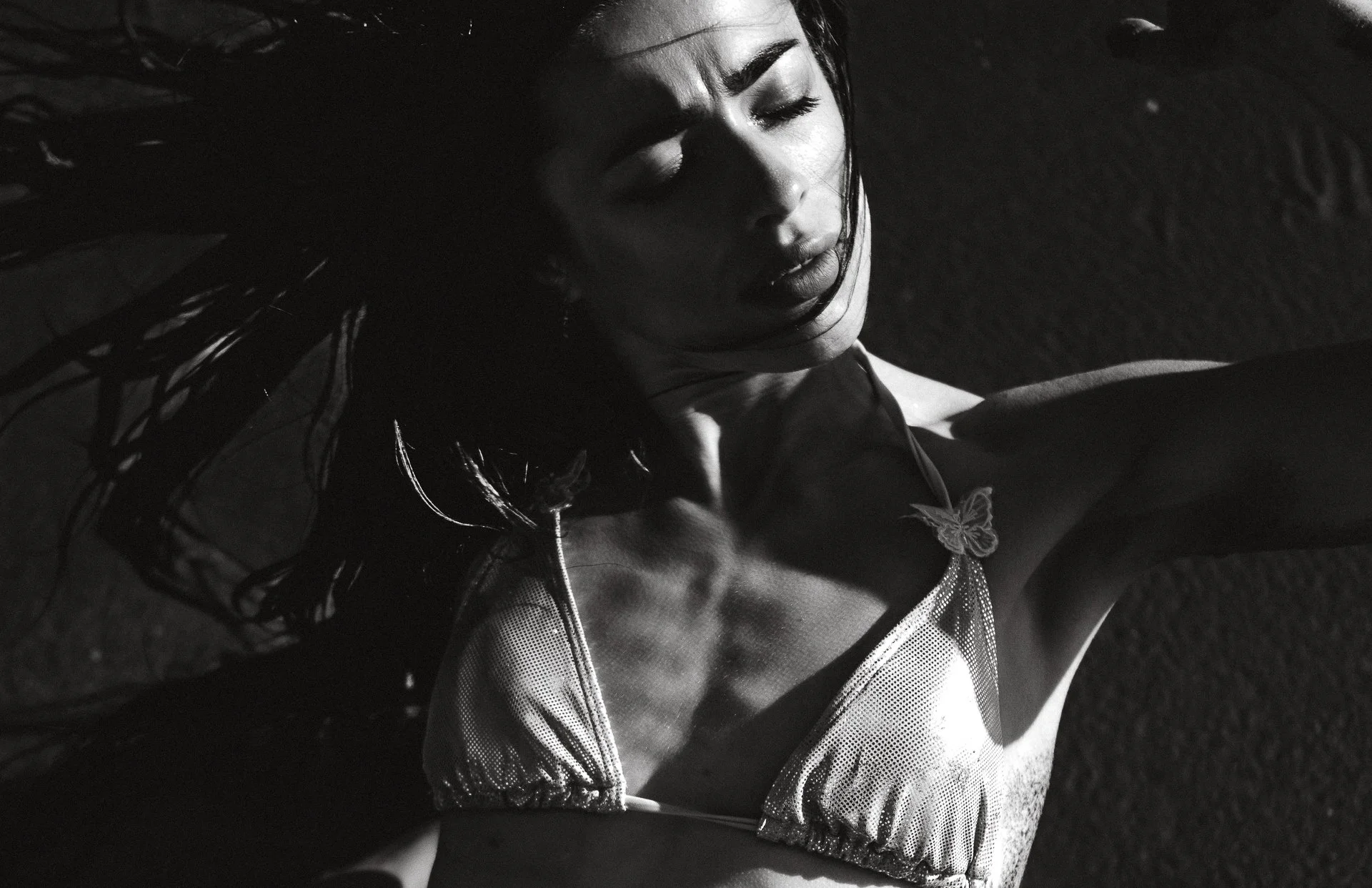 Black and white photo of a woman with her eyes closed, wearing a bikini with a butterfly ornament, lying on the ground with her hair spread out.