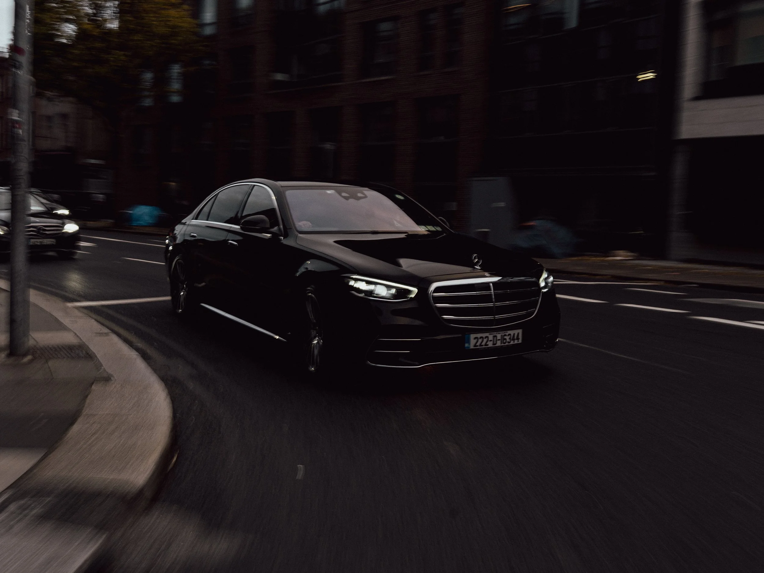 Black luxury sedan driving on city street at dusk, dark buildings and other cars in the background.