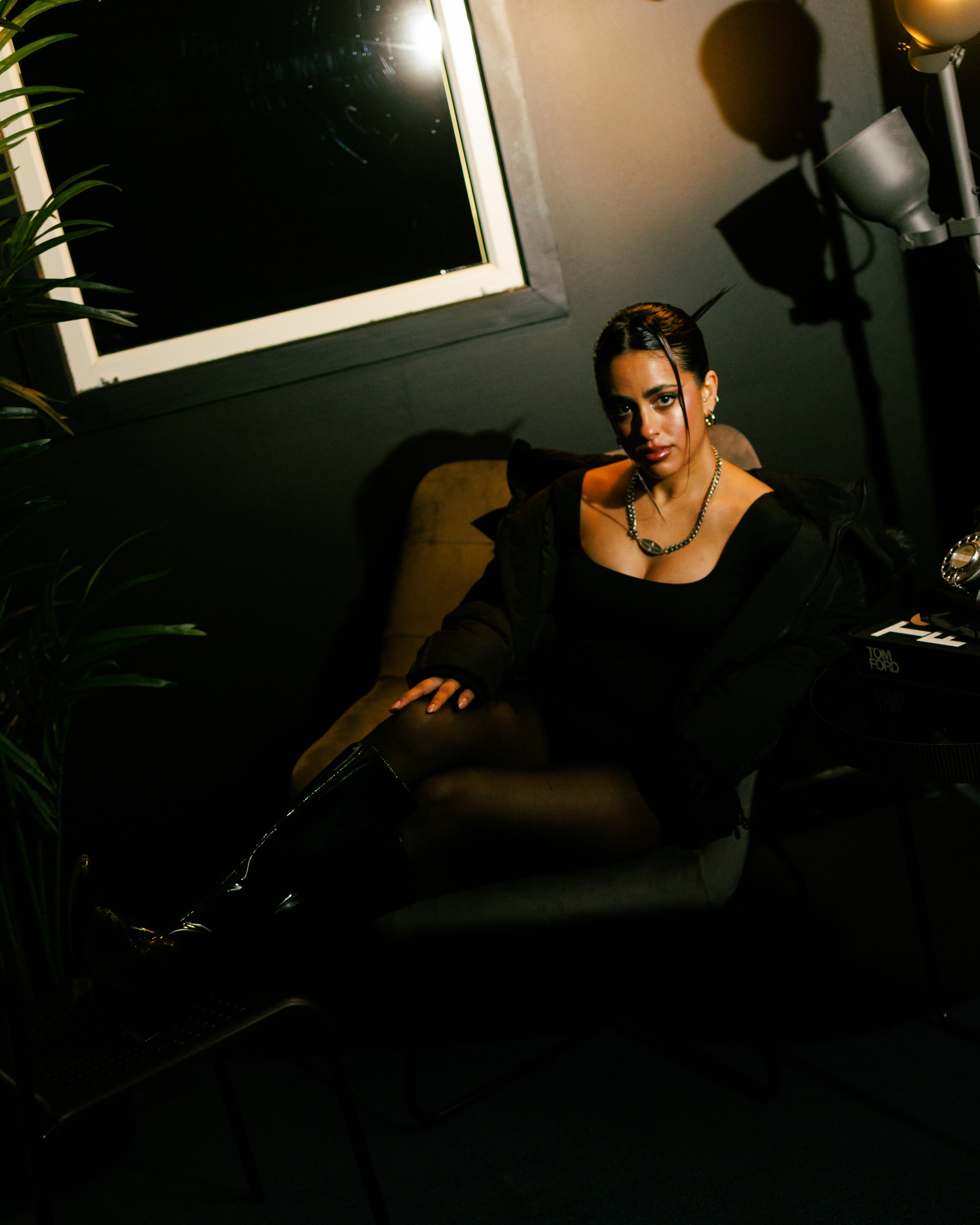 A woman sitting on a brown couch in a dimly lit room, wearing black attire, with dark hair styled back, jewelry, and knee-high boots.