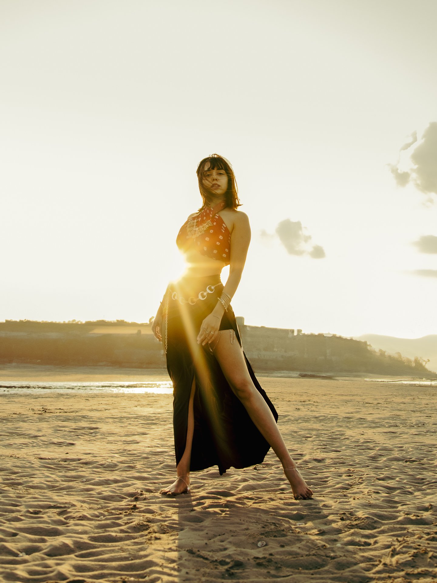 A woman in a black skirt with a high slit and a red top standing on the beach during sunset, with the sun shining behind her creating a glow and lens flare.