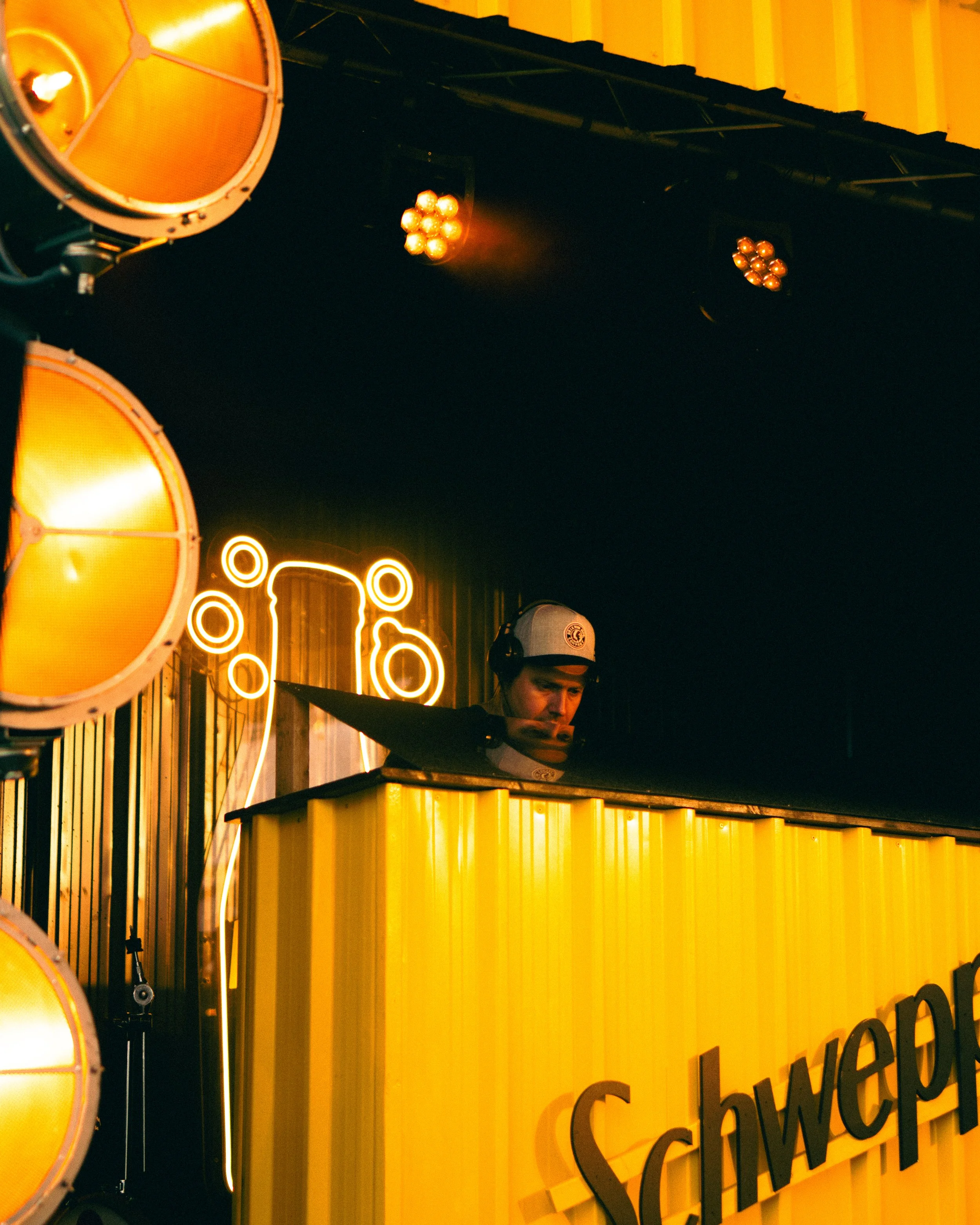 A DJ wearing a white cap and headphones performing behind a yellow booth with the word 'Schwep' visible, illuminated by yellow stage lights with neon circular decorations in the background.