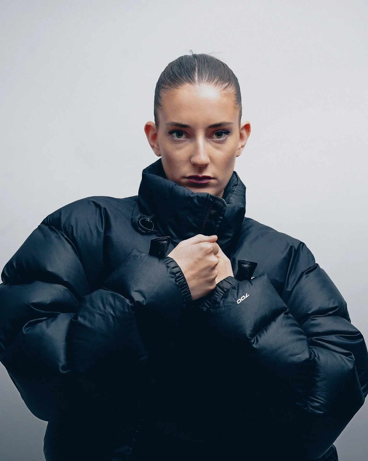 A young woman with a serious expression, wearing a black puffer jacket with a high collar, standing against a plain background.
