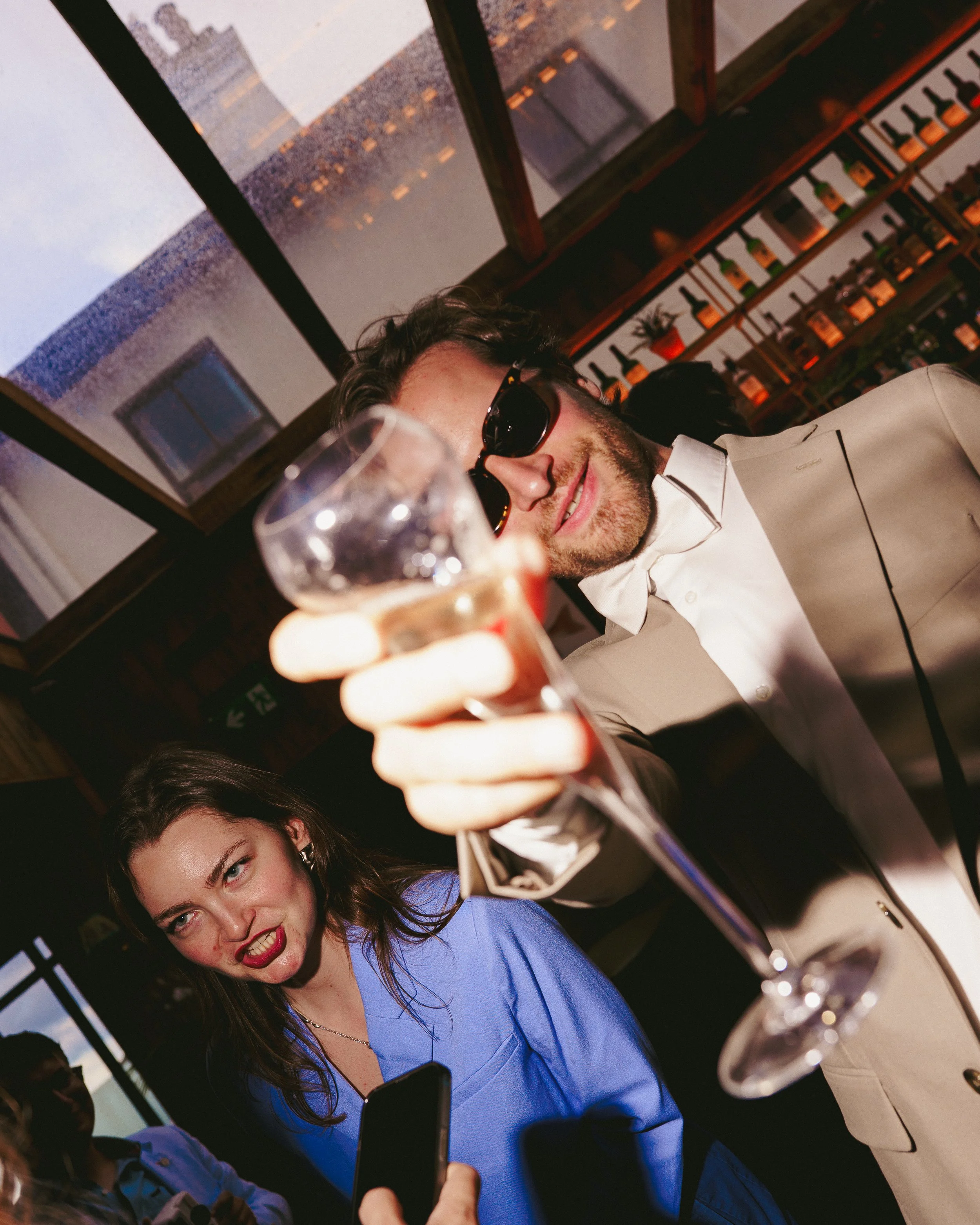 Young man in a tuxedo and sunglasses holding a glass of champagne, smiling, at a party or celebration with a woman in a blue dress and other guests in the background.