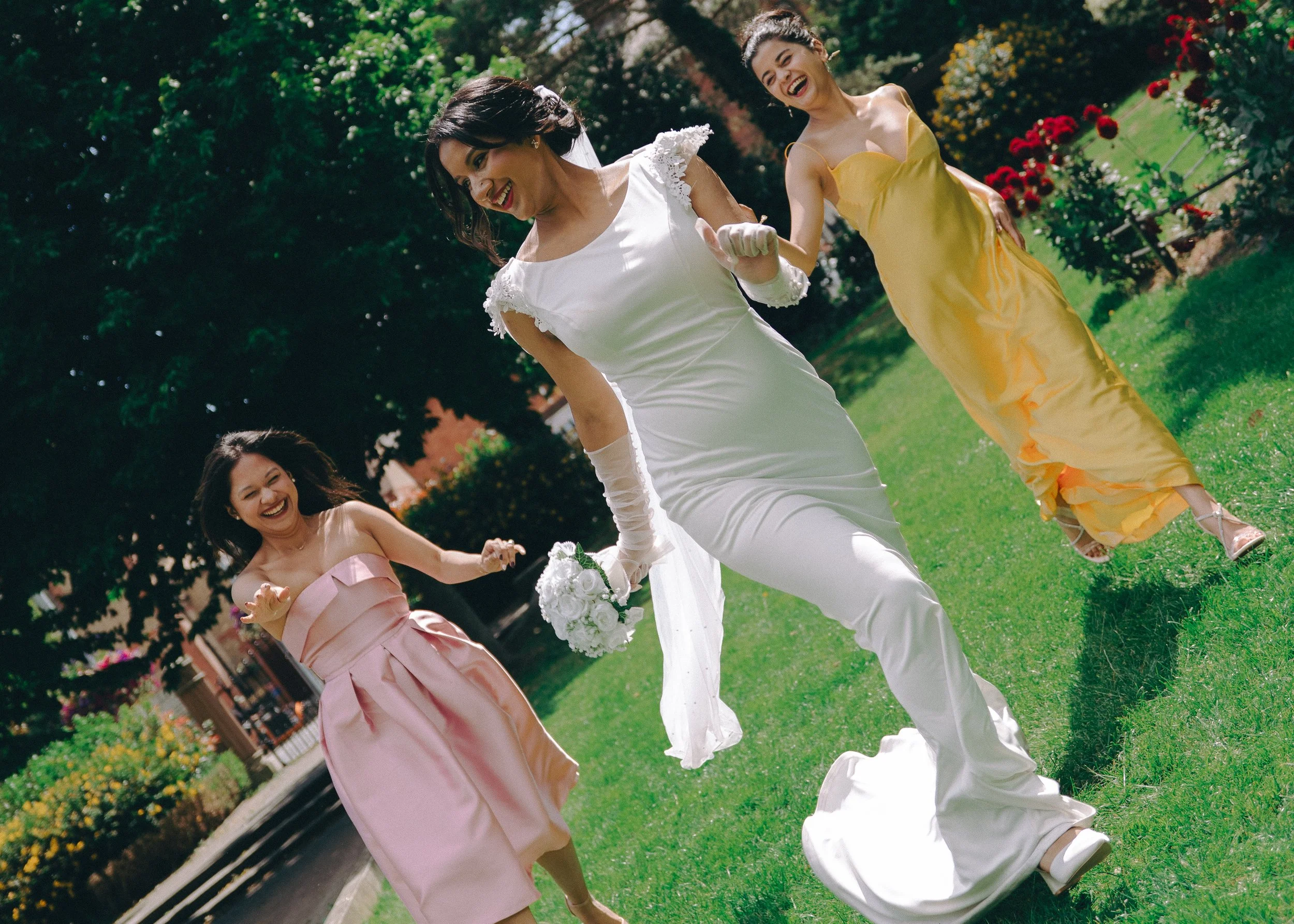 Three women in colorful dresses playing and laughing on a grassy lawn surrounded by trees and flowers, enjoying a sunny day.