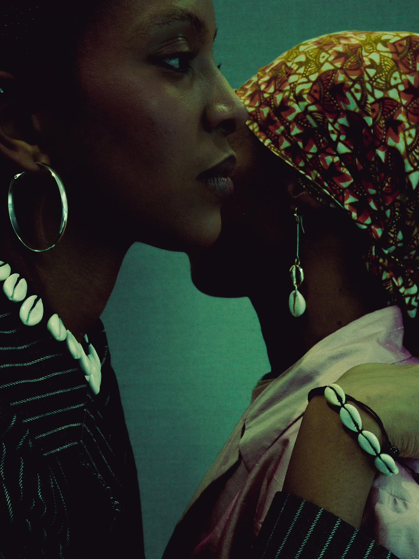 Close-up of two women with dark skin, wearing colorful earrings and shell jewelry, in profile view, with one woman wearing a patterned headwrap.