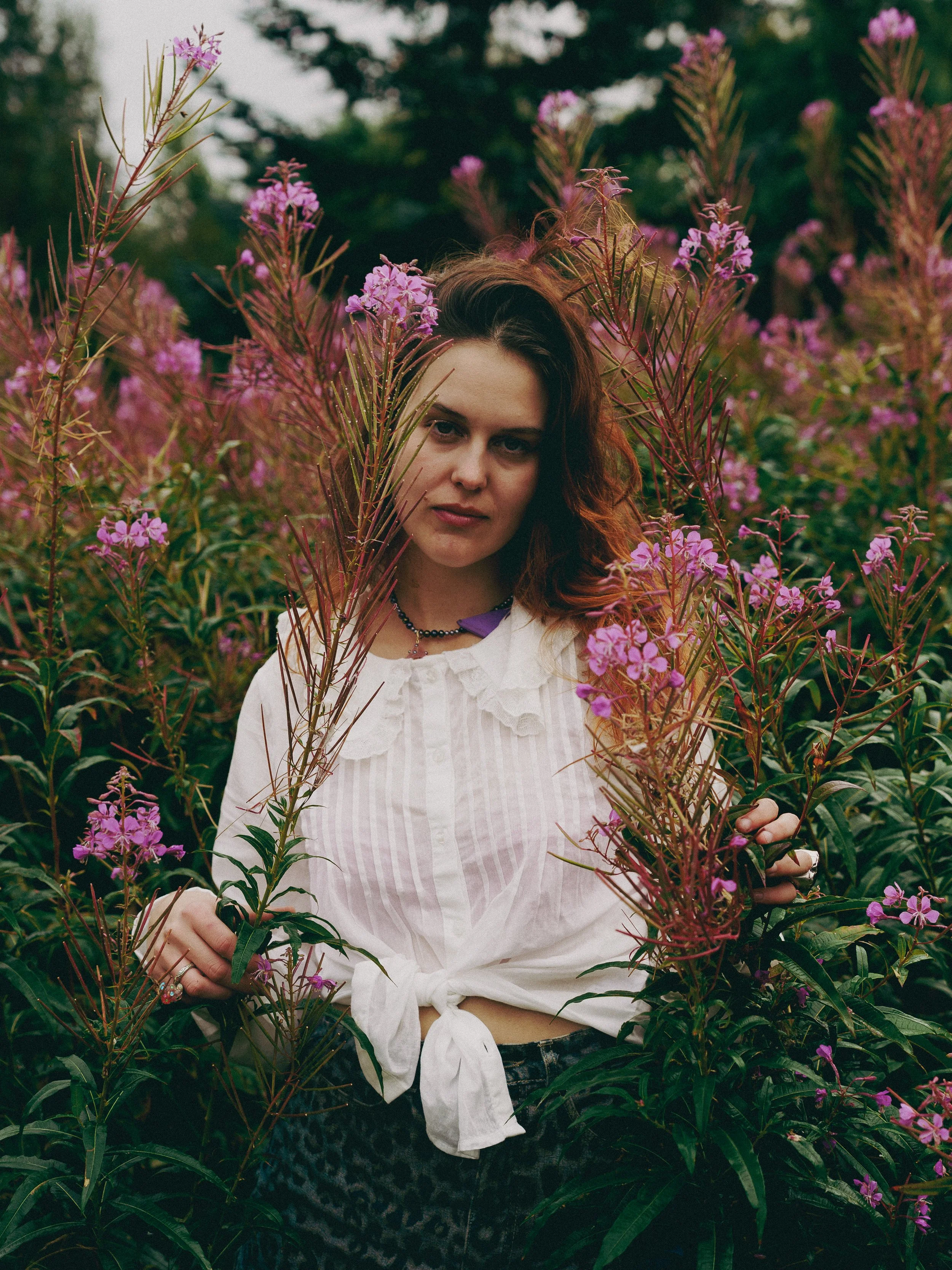 Woman with wavy reddish-brown hair standing among tall pink and purple flowers in a garden or natural setting.
