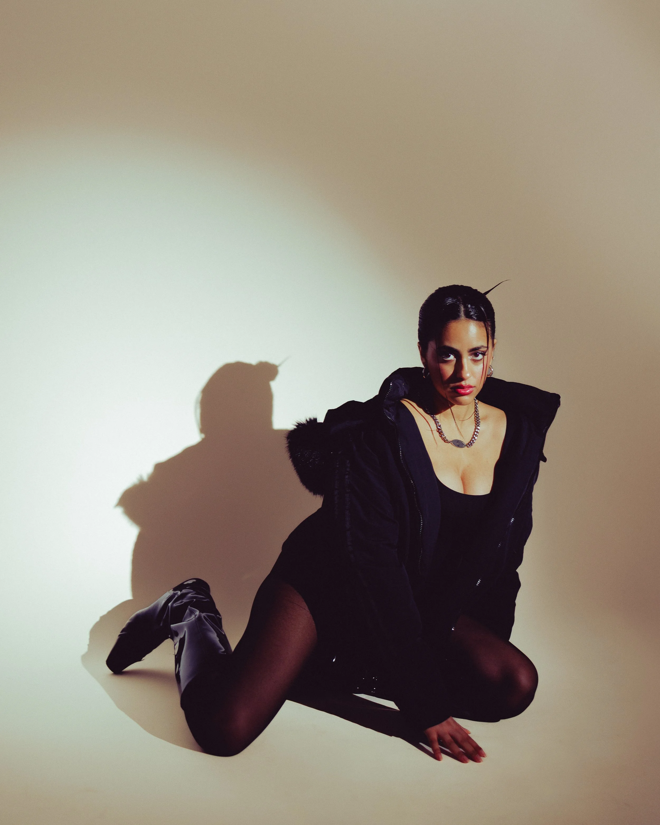 A woman kneeling on the floor, wearing black clothing and boots, with a serious expression, against a neutral background and casting a shadow.