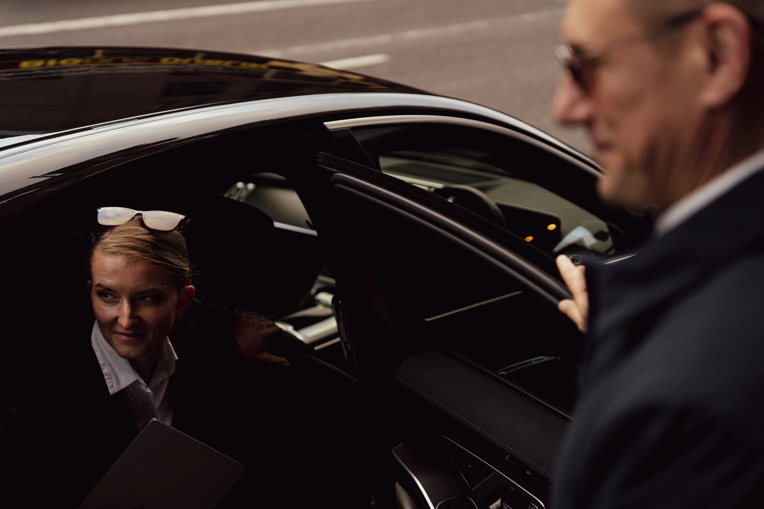 A woman sitting inside a black car with sunglasses on her head, looking out of the window at a man standing outside the car, holding the door.