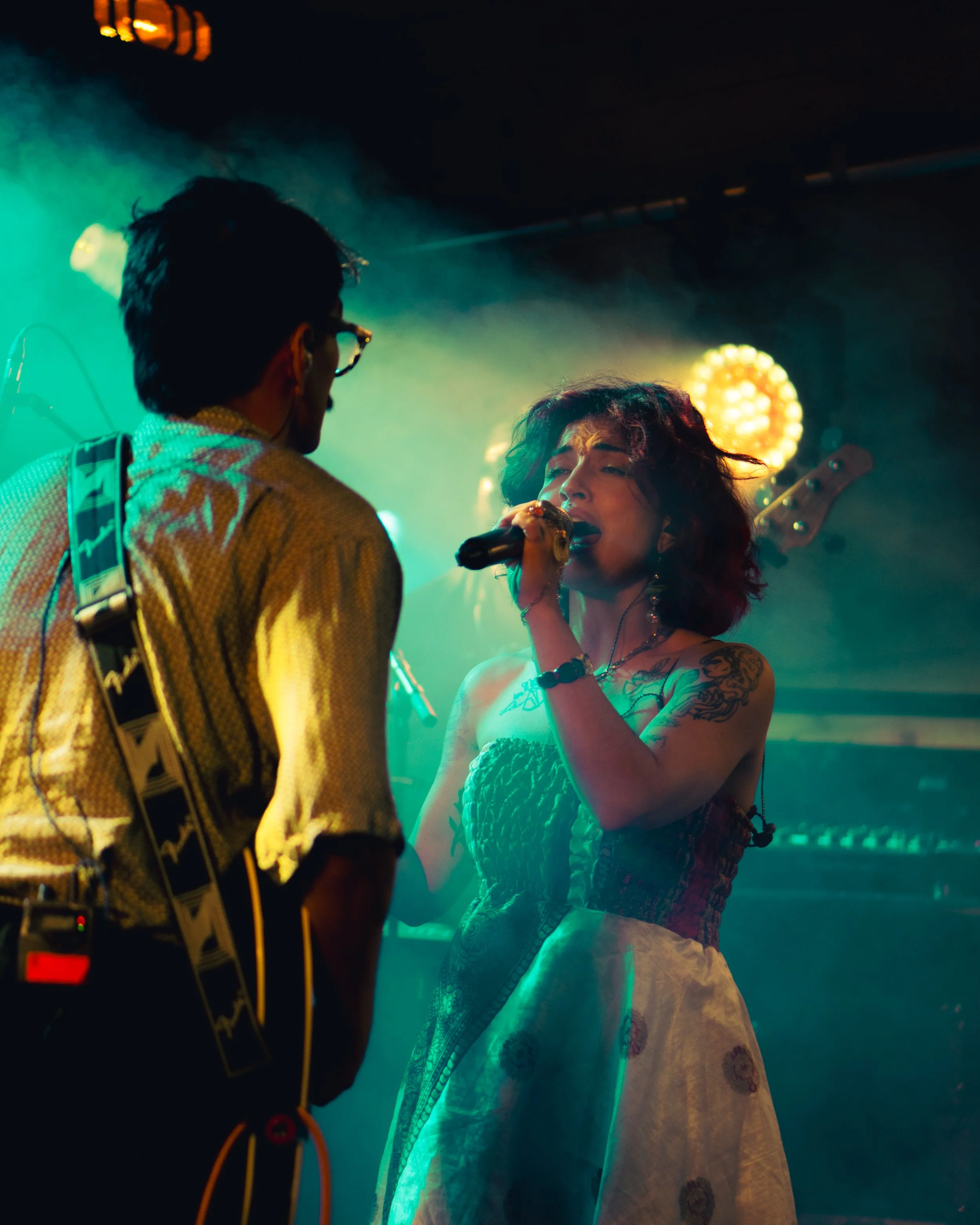 A woman singing passionately into a microphone on stage with a guitarist facing her, illuminated by colorful stage lights.