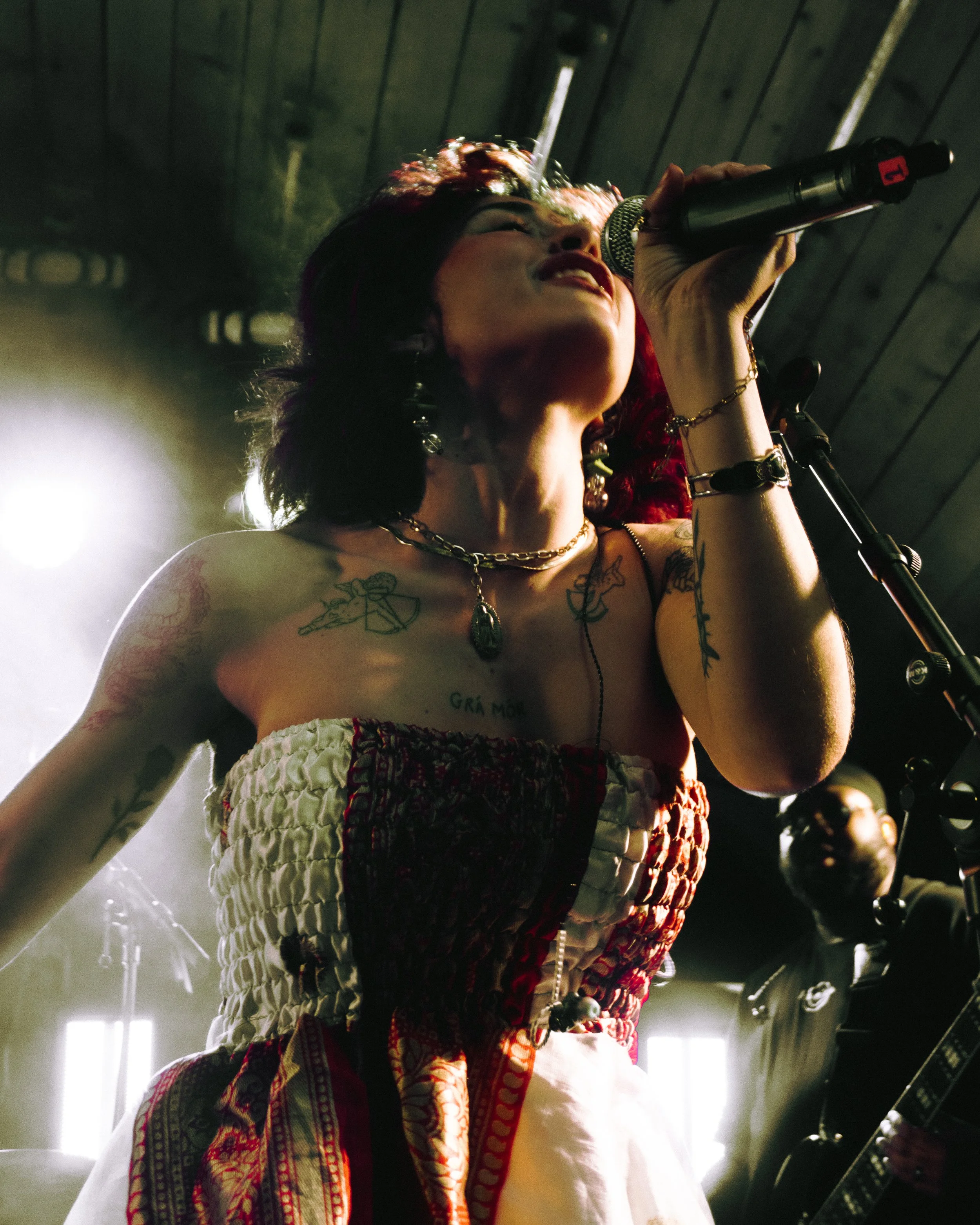A woman singing into a microphone during a live performance, with tattoos on her chest and shoulders, wearing jewelry and a strapless dress.