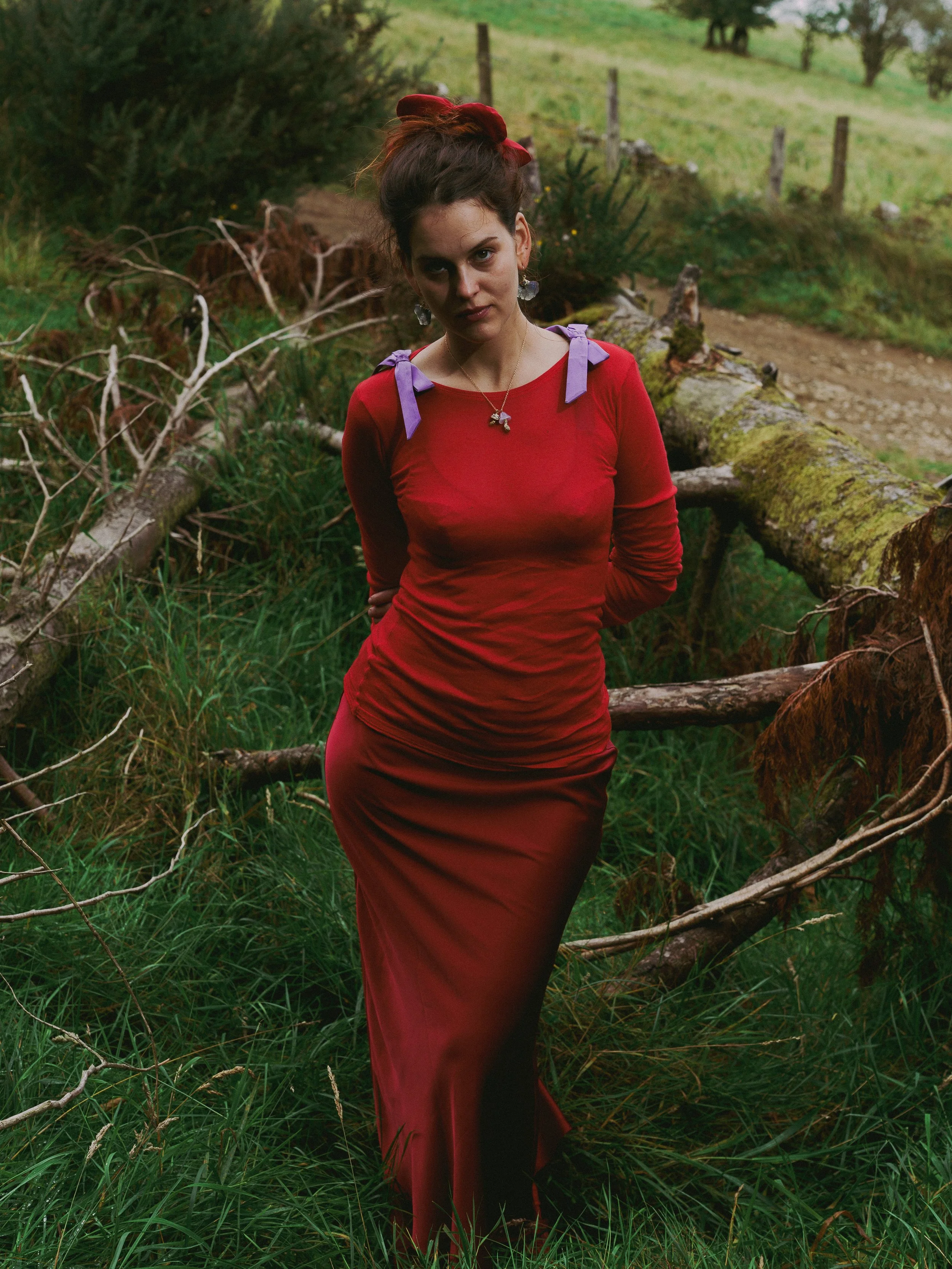 A woman with dark hair tied with a red ribbon, wearing a long red dress with purple bows on the shoulders, standing outdoors in a grassy area with fallen trees and a dirt path in the background.