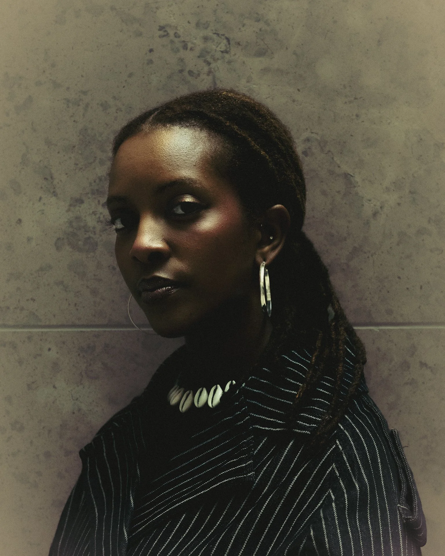 A woman with dark skin, long braided hair, and bold makeup poses against a textured wall, wearing large hoop earrings, a necklace, and a black striped outfit.