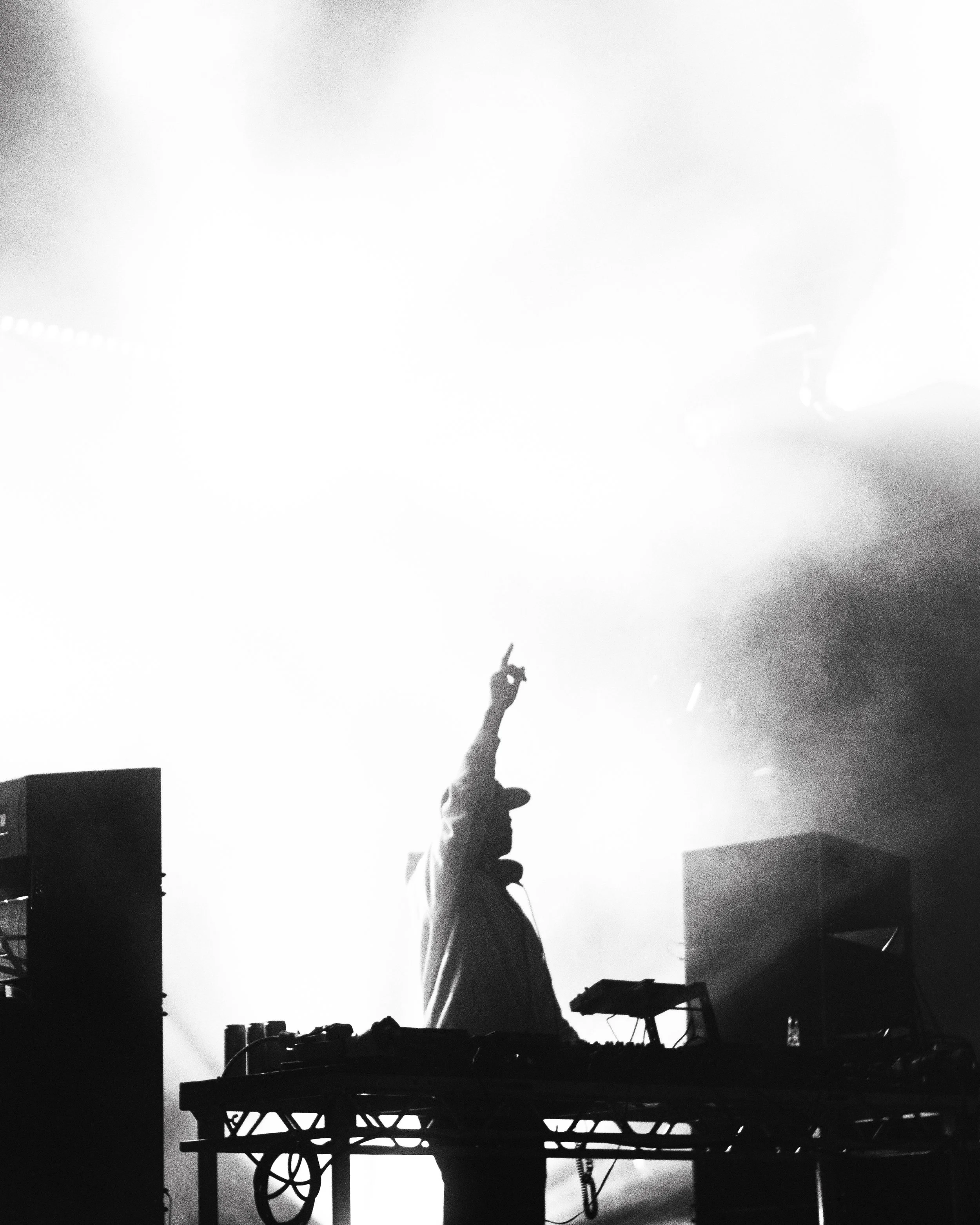 A DJ performs on stage in front of bright lights, raising one hand with the index finger pointing up, with equipment and speakers around.