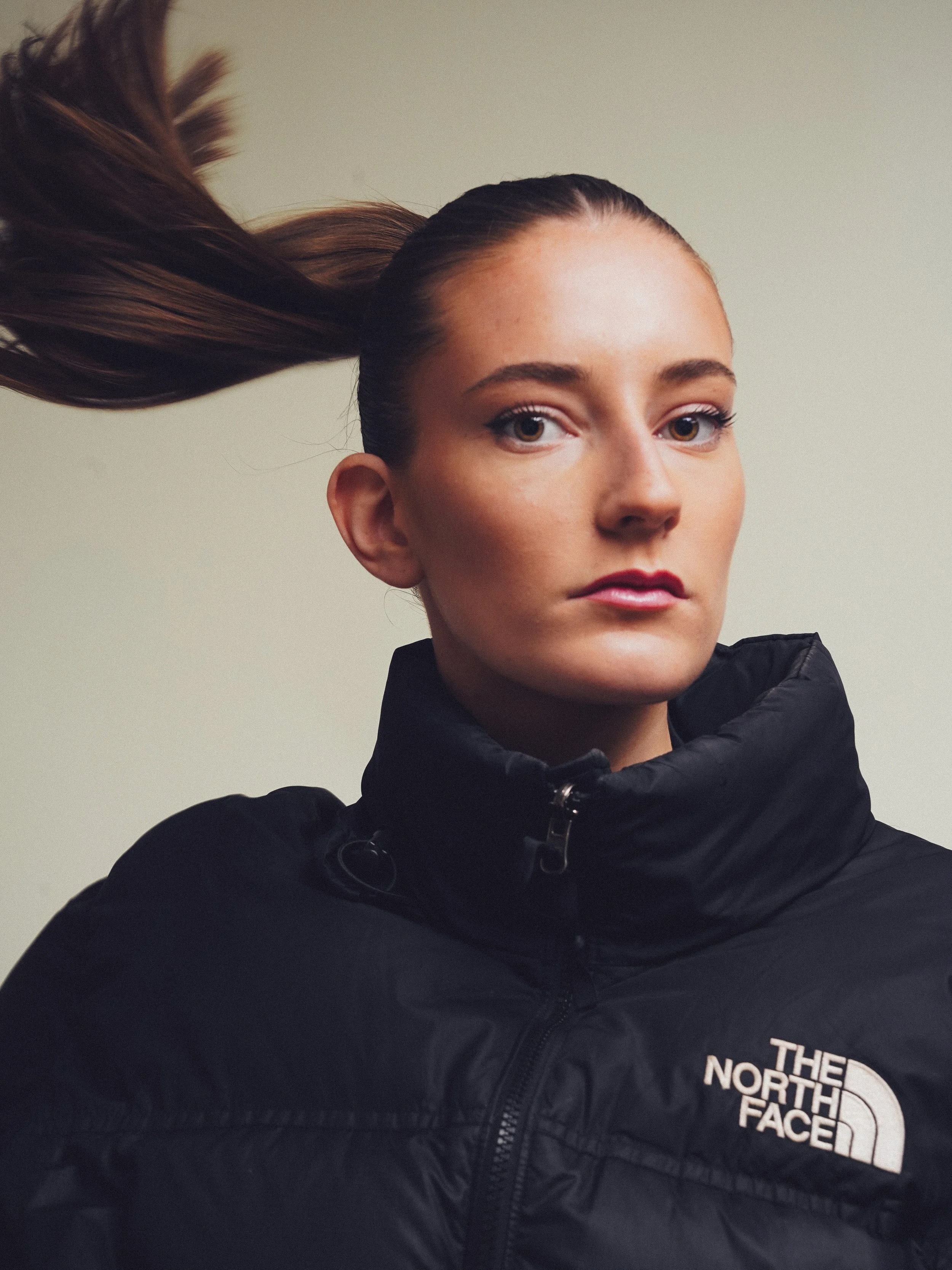 A woman with long brown hair, wearing a black North Face jacket, looking directly at the camera against a neutral background.