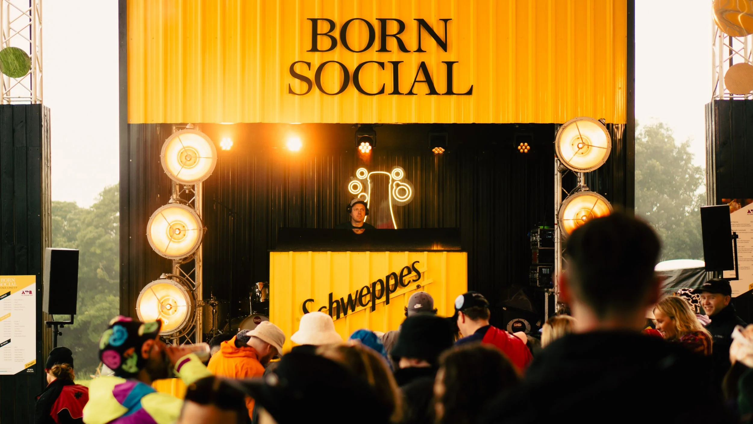 A DJ performs on a stage with a large yellow sign that says 'Born Social' and a smaller sign with the Schweppes logo. The stage is lit with bright yellow lights, and there is a crowd watching. Trees are visible in the background.
