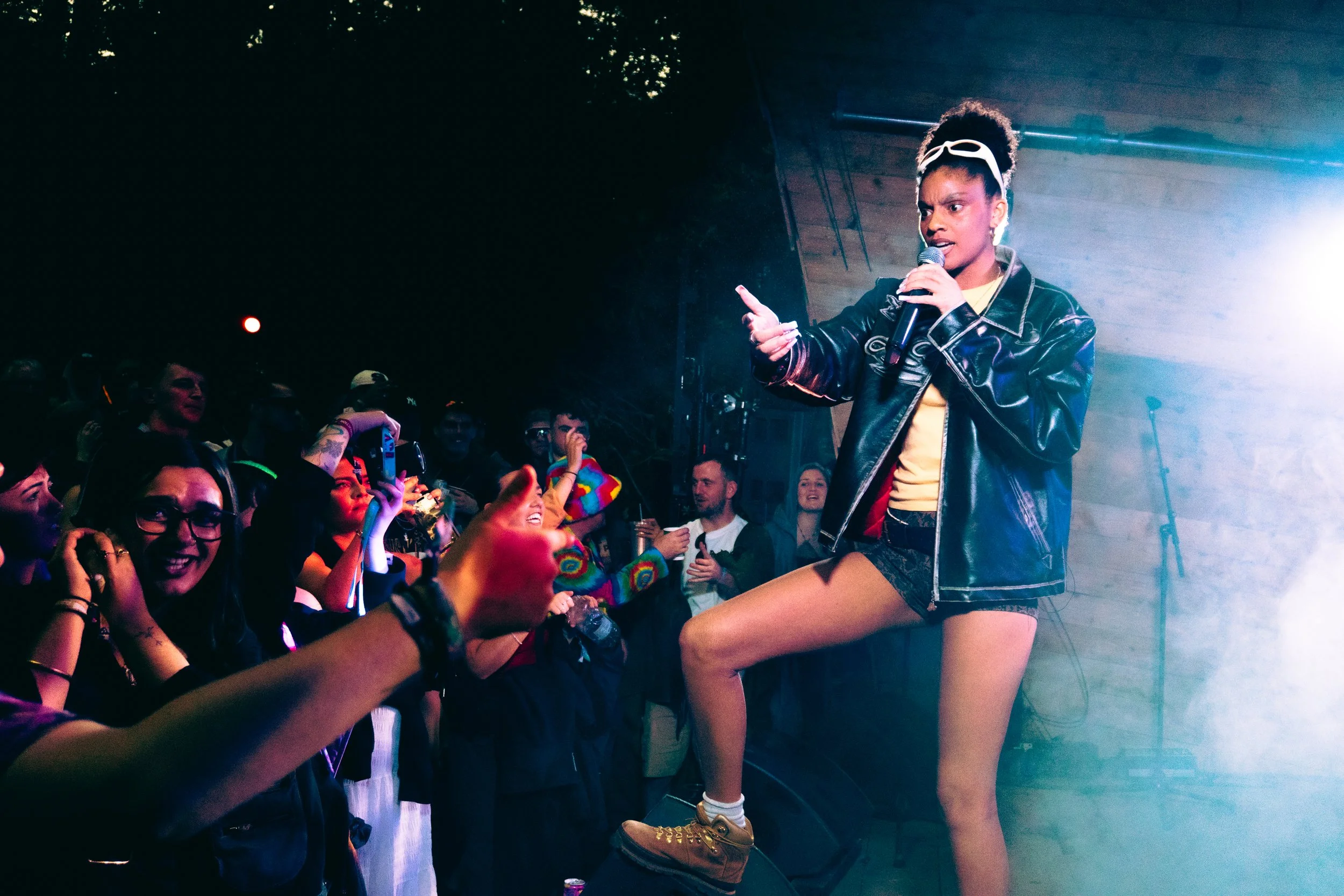 Performer singing into a microphone on stage, dressed in a black leather jacket, yellow top, shorts, and brown boots, with a crowd cheering in front of her at a concert.
