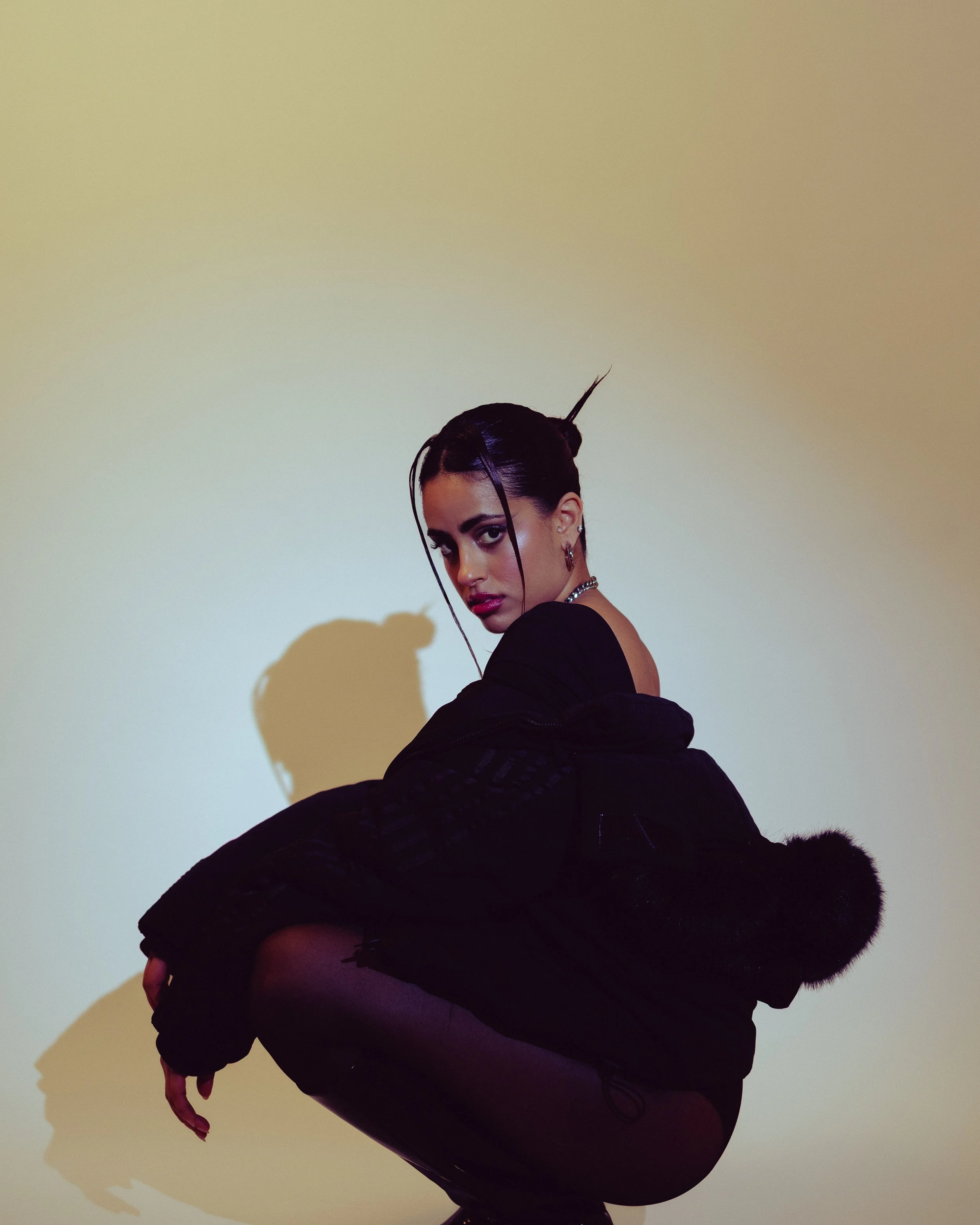A woman with dark hair styled in a sleek updo with a hairpin, wearing a black outfit with a fur hood, squatting against a plain light-colored background, casting a shadow on the wall.