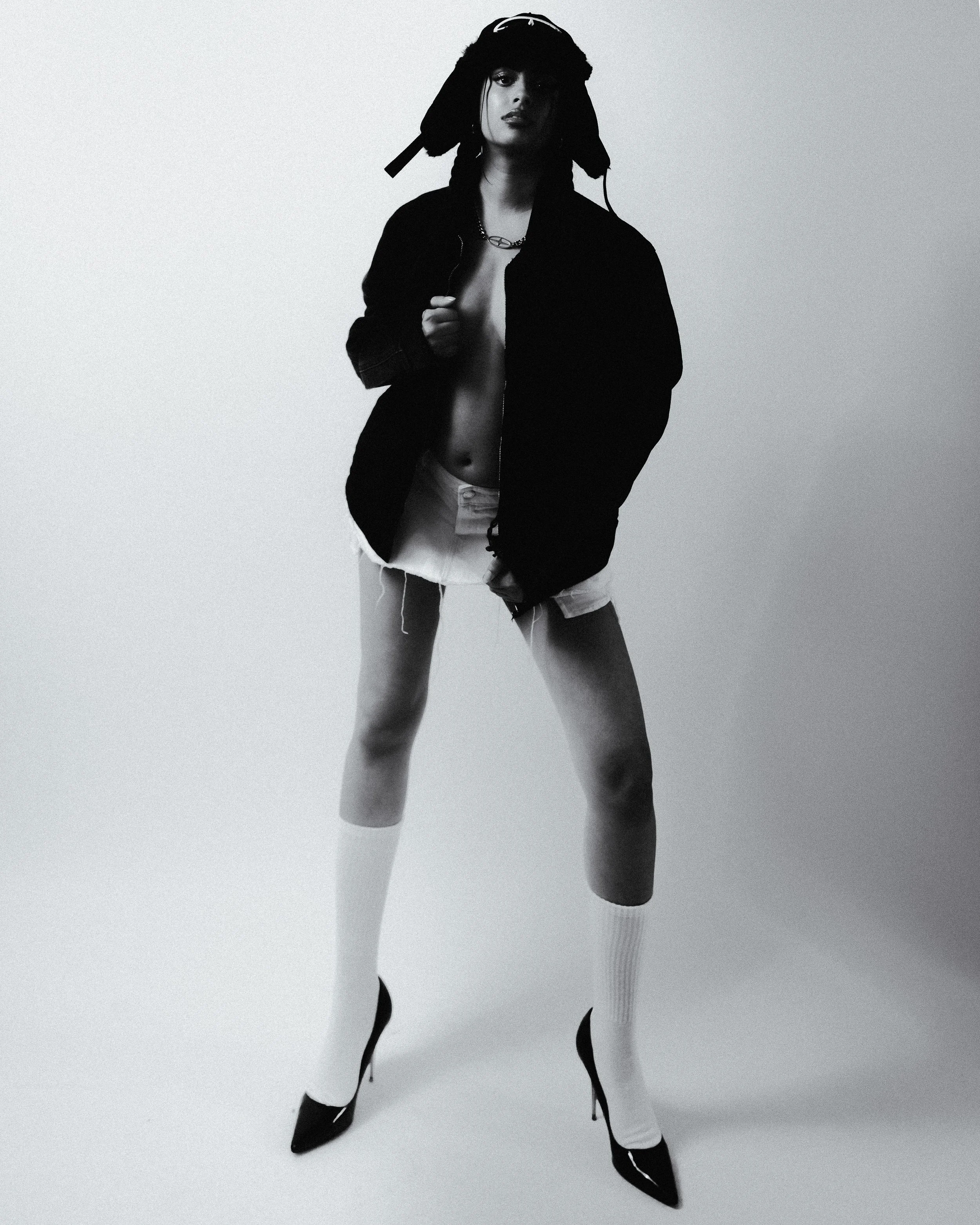 A woman standing with one leg in front of the other against a plain background. She is wearing high heels, knee-high socks, a jacket, and a hat, with her shirt partially open revealing her chest.