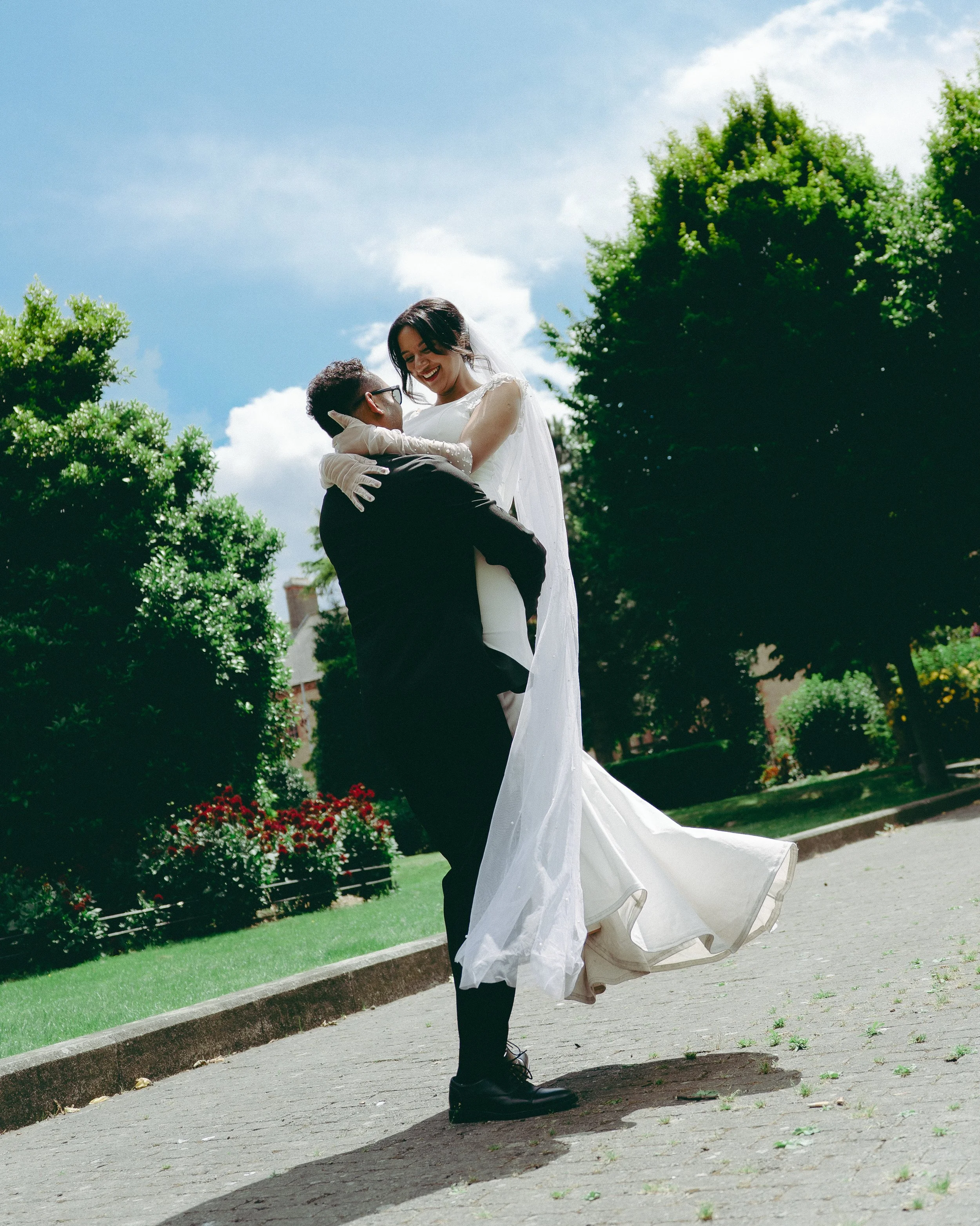 A man in a black suit lifting a woman in a white wedding dress outdoors, surrounded by green trees and flowers, under a partly cloudy sky.