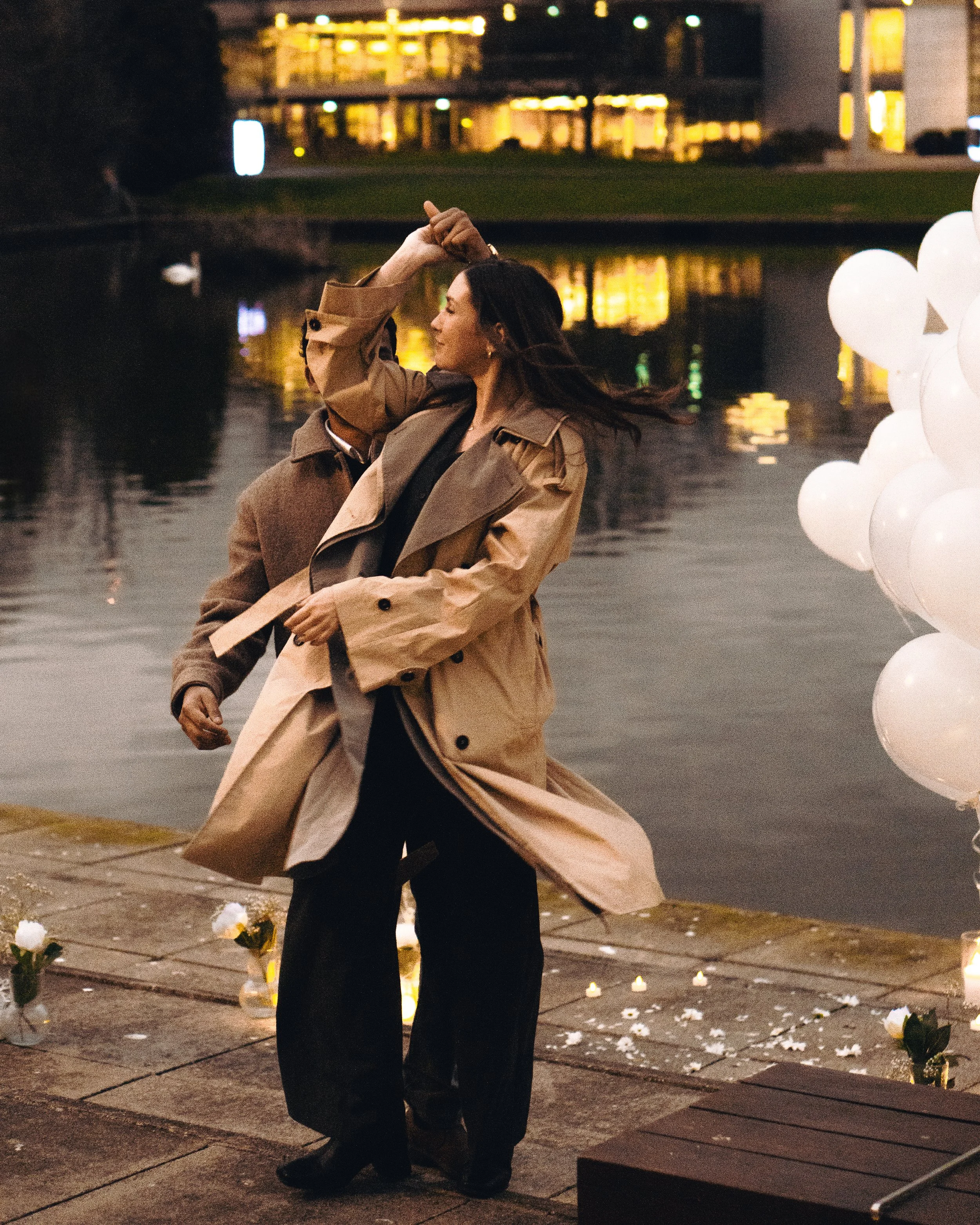 A woman in a beige trench coat dancing by a waterfront at night, with candles and white balloons nearby and a building with lit windows in the background.