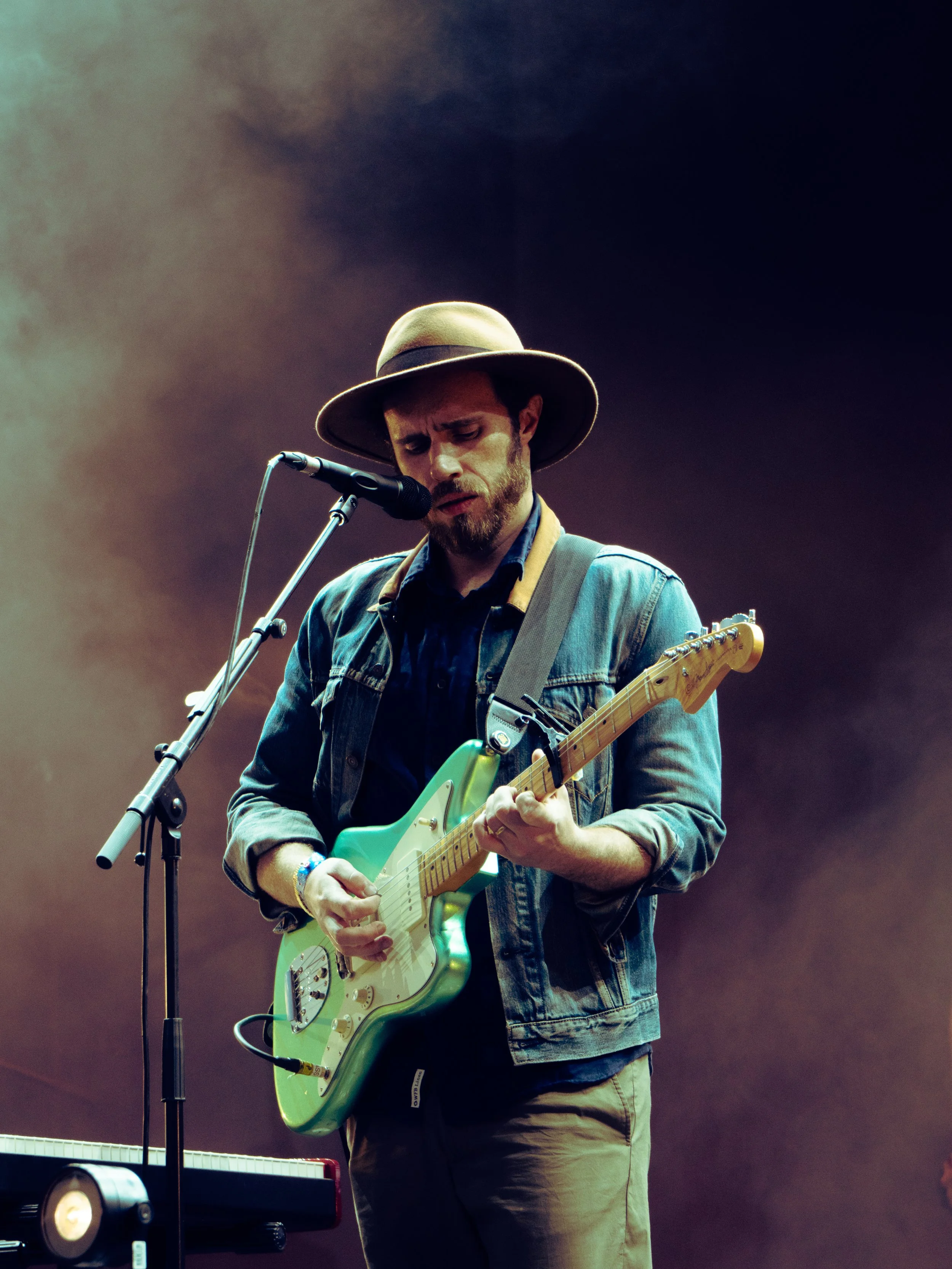 A man wearing a wide-brimmed hat and denim jacket playing an electric guitar on stage with smoke in the background.