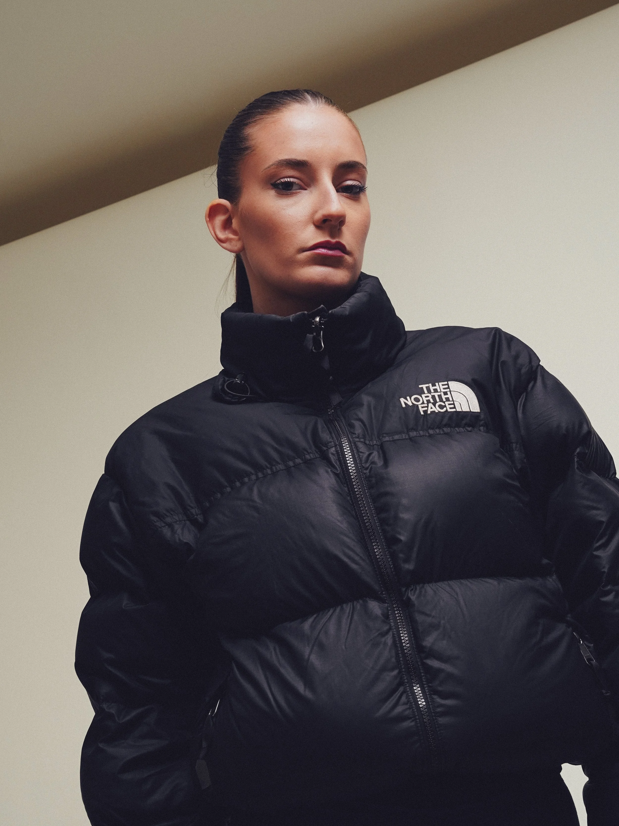 A woman with slicked-back hair wearing a black The North Face puffer jacket looking confidently at the camera against a plain light-colored wall.