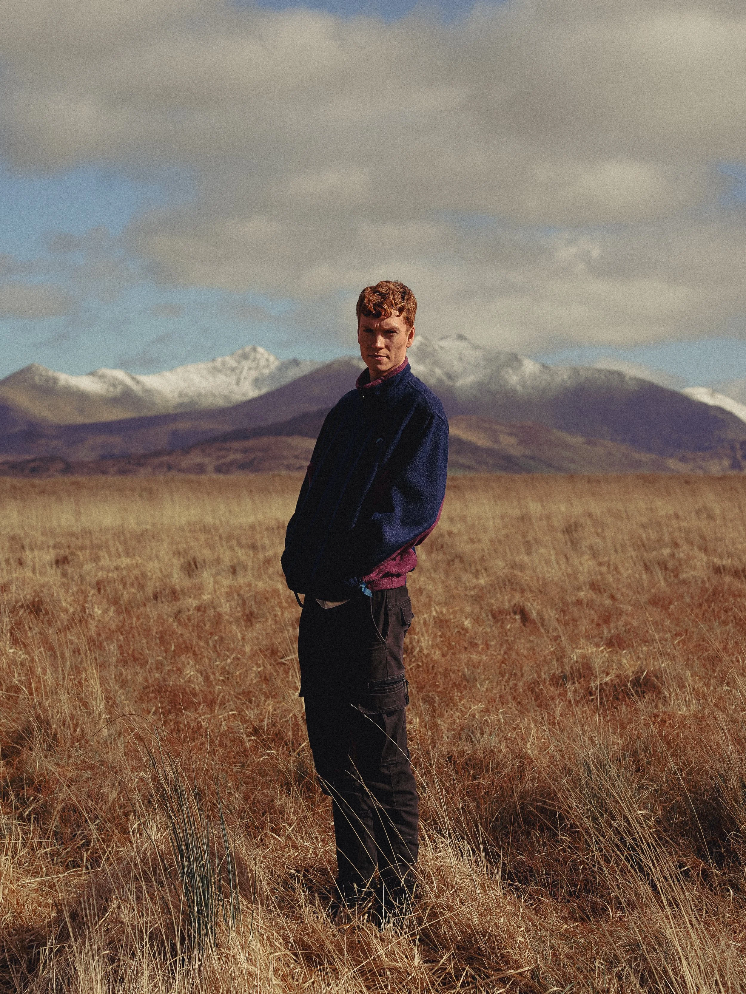 A young man standing in a vast field with tall grass, mountains with snow on their peaks in the background, and cloudy skies overhead.
